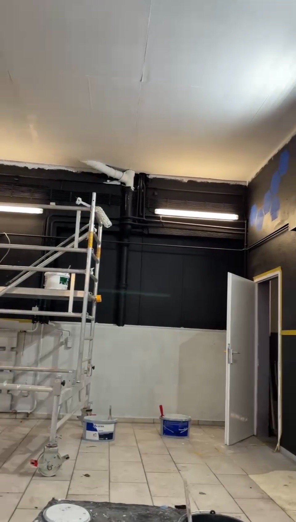 Wnętrze w trakcie malowania: rusztowanie, puszki z farbą, pomalowany sufit i ściany w kolorze czarnym i białym. Widoczne instalacje pod sufitem.