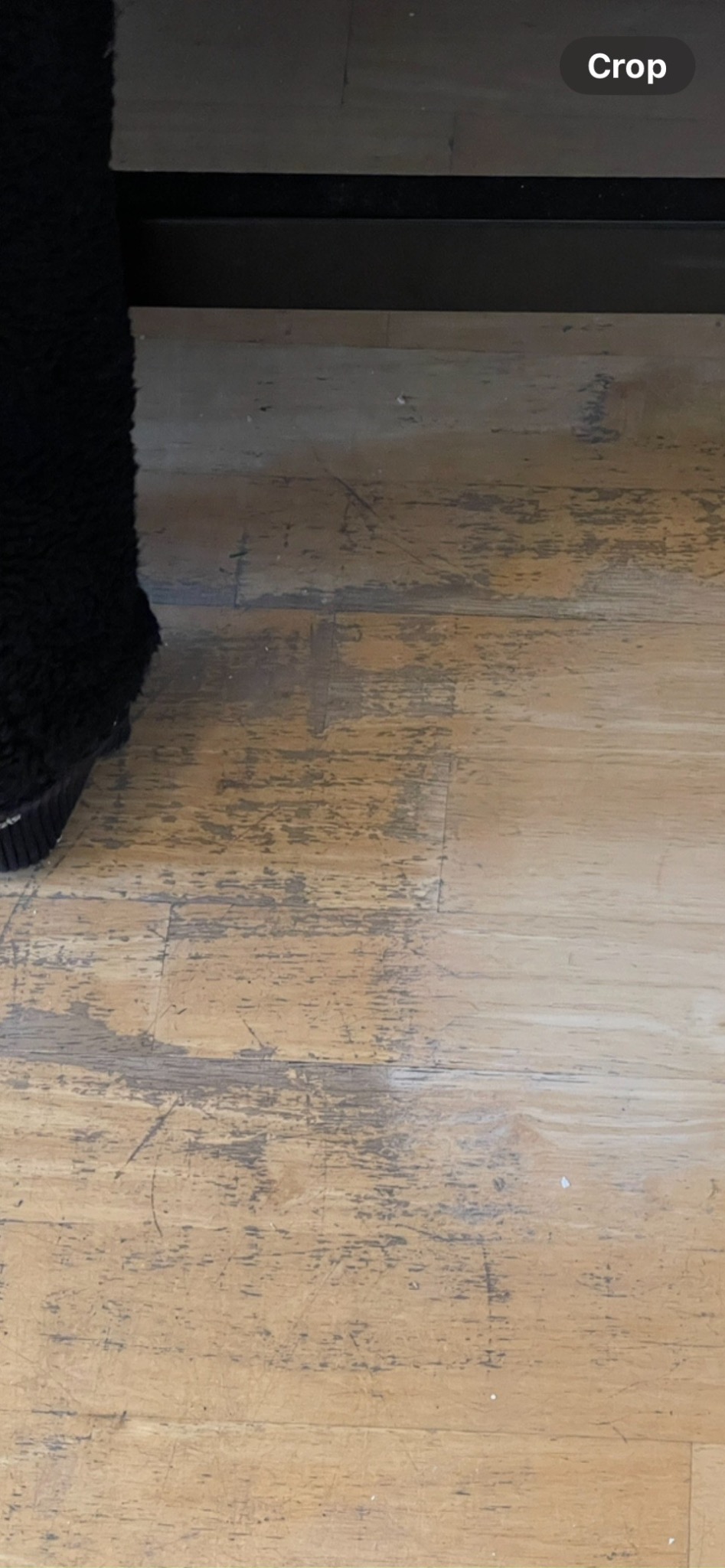 Jasne panele podłogowe z widocznymi śladami użytkowania i zarysowaniami, fragment czarnego mebla w lewym górnym rogu kadru.
