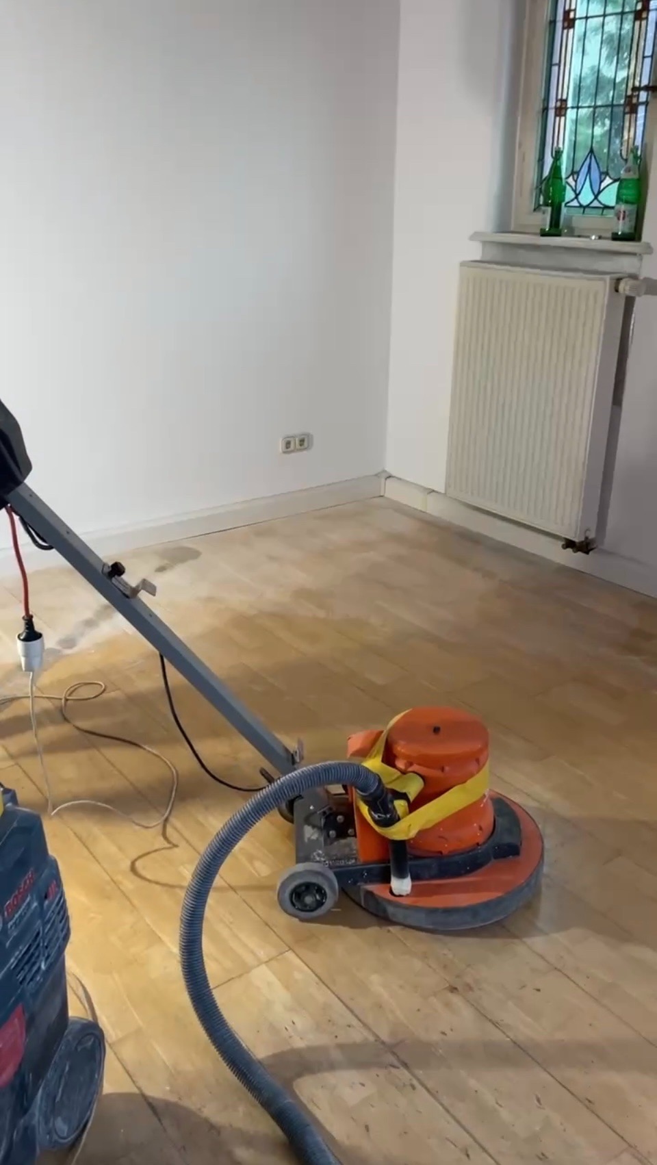 Szlifowanie parkietu maszyną w jasnym pokoju z oknem witrażowym. Widoczny fragment odkurzacza przemysłowego. Podłoga częściowo wyszlifowana, ukazująca jaśniejszy odcień drewna.