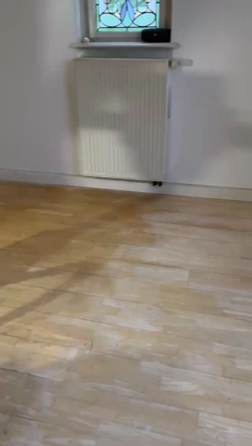 Jasne panele podłogowe z widocznym grzejnikiem i oknem witrażowym w tle. Minimalistyczne wnętrze po remoncie, ukazujące efekt wykończenia podłogi.