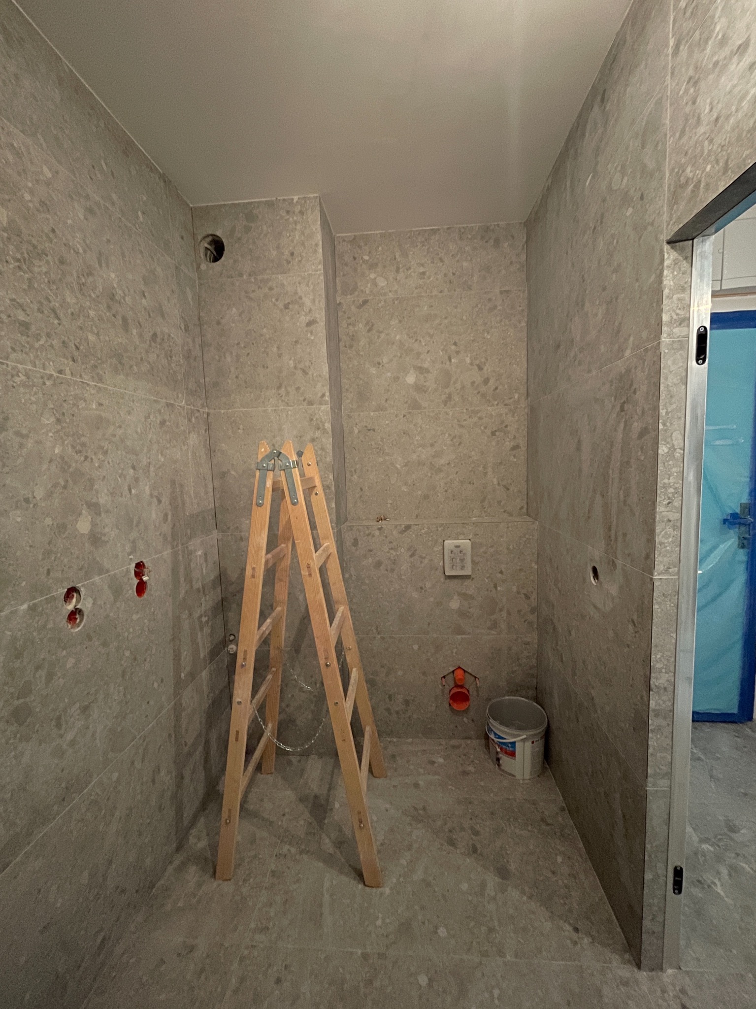 Łazienka w trakcie wykańczania: szare płytki imitujące kamień na ścianach i podłodze, drewniana drabina, widoczne instalacje i przygotowane otwory. Drzwi zabezpieczone folią.
