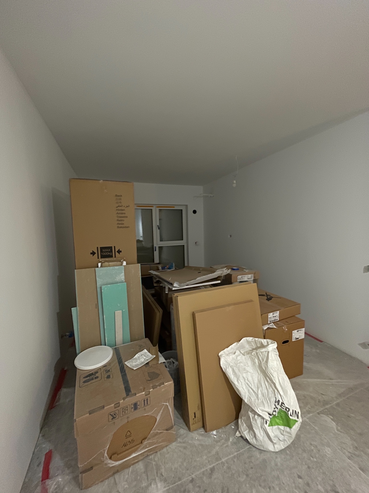 Pokój w trakcie remontu: kartony, płyty gipsowe, worki na gruz, folia ochronna na podłodze. Widoczne okno i surowe ściany. Przygotowanie do prac wykończeniowych.