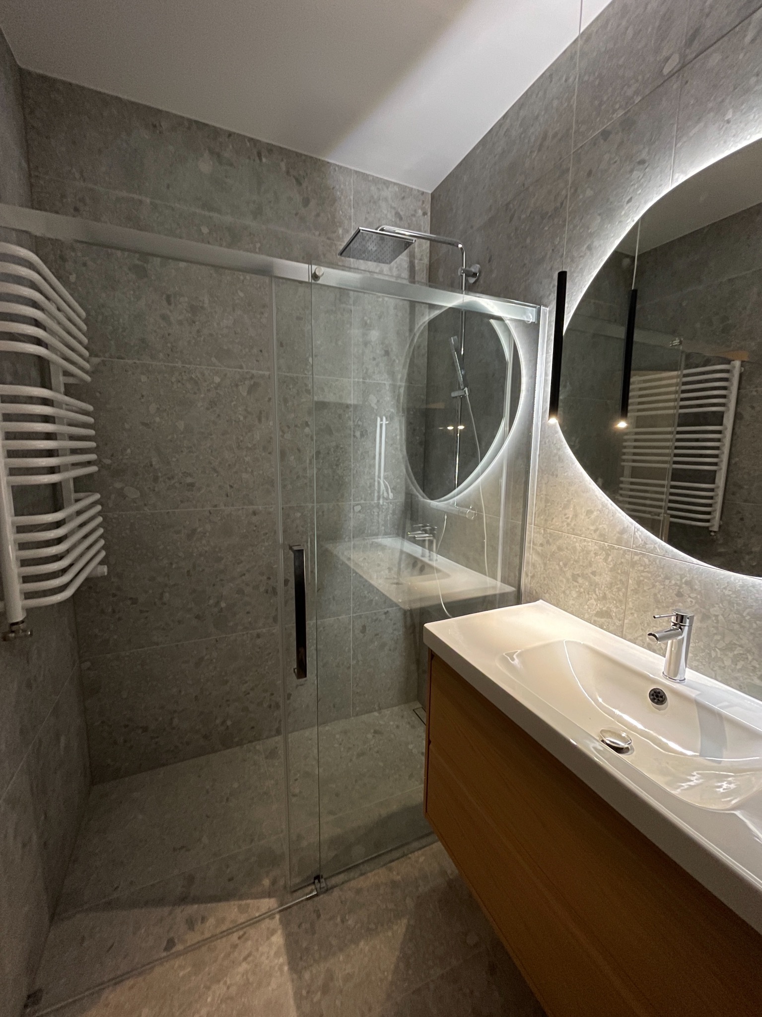 Nowoczesna łazienka z kabiną prysznicową typu walk-in, umywalką z szafką z drewnopodobnym dekorem i okrągłym lustrem z podświetleniem LED.