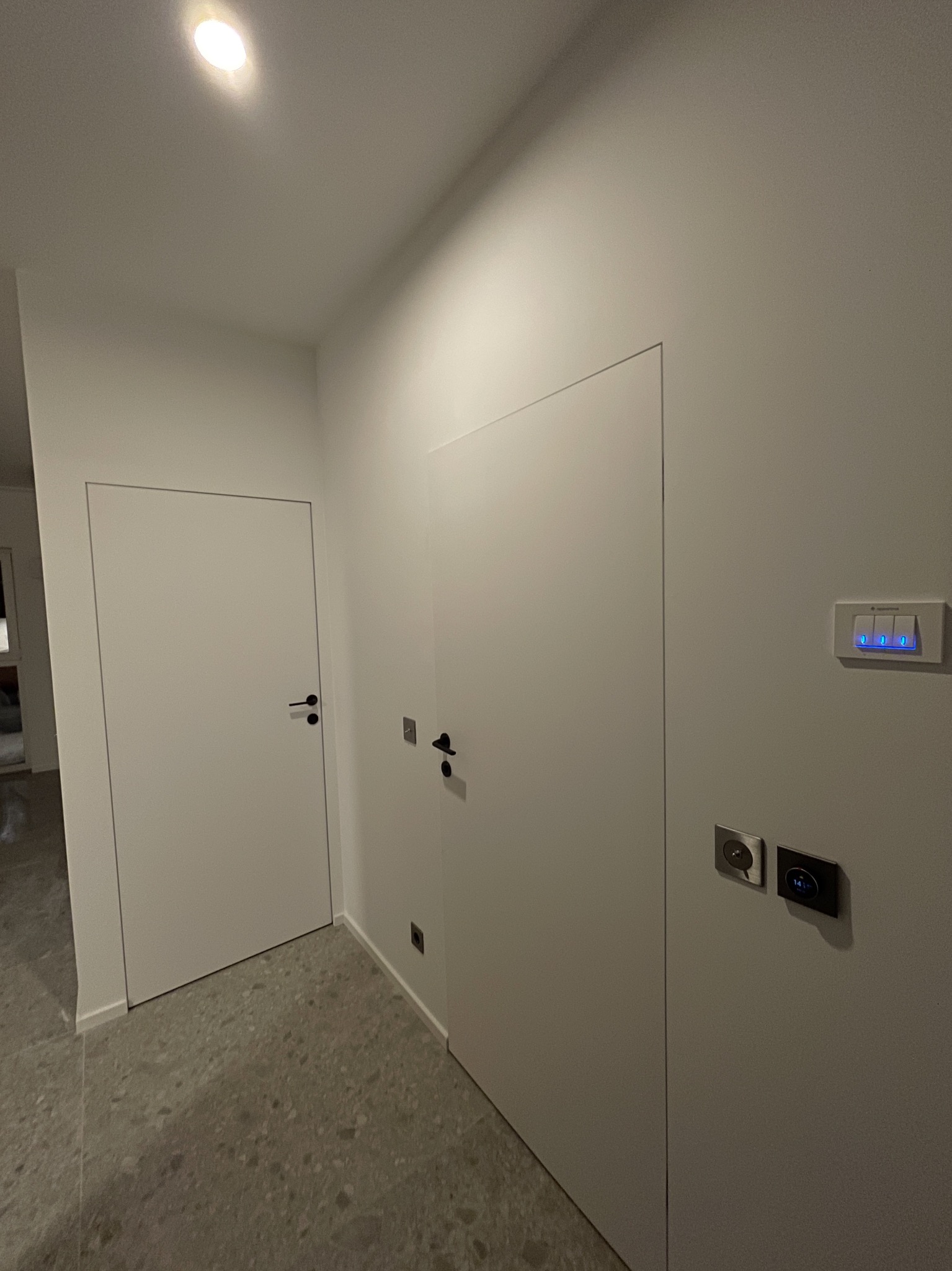 Minimalistyczny korytarz z dwiema białymi, ukrytymi drzwiami i szarą posadzką lastryko. Na ścianie włączniki z niebieskim podświetleniem i gniazdka.