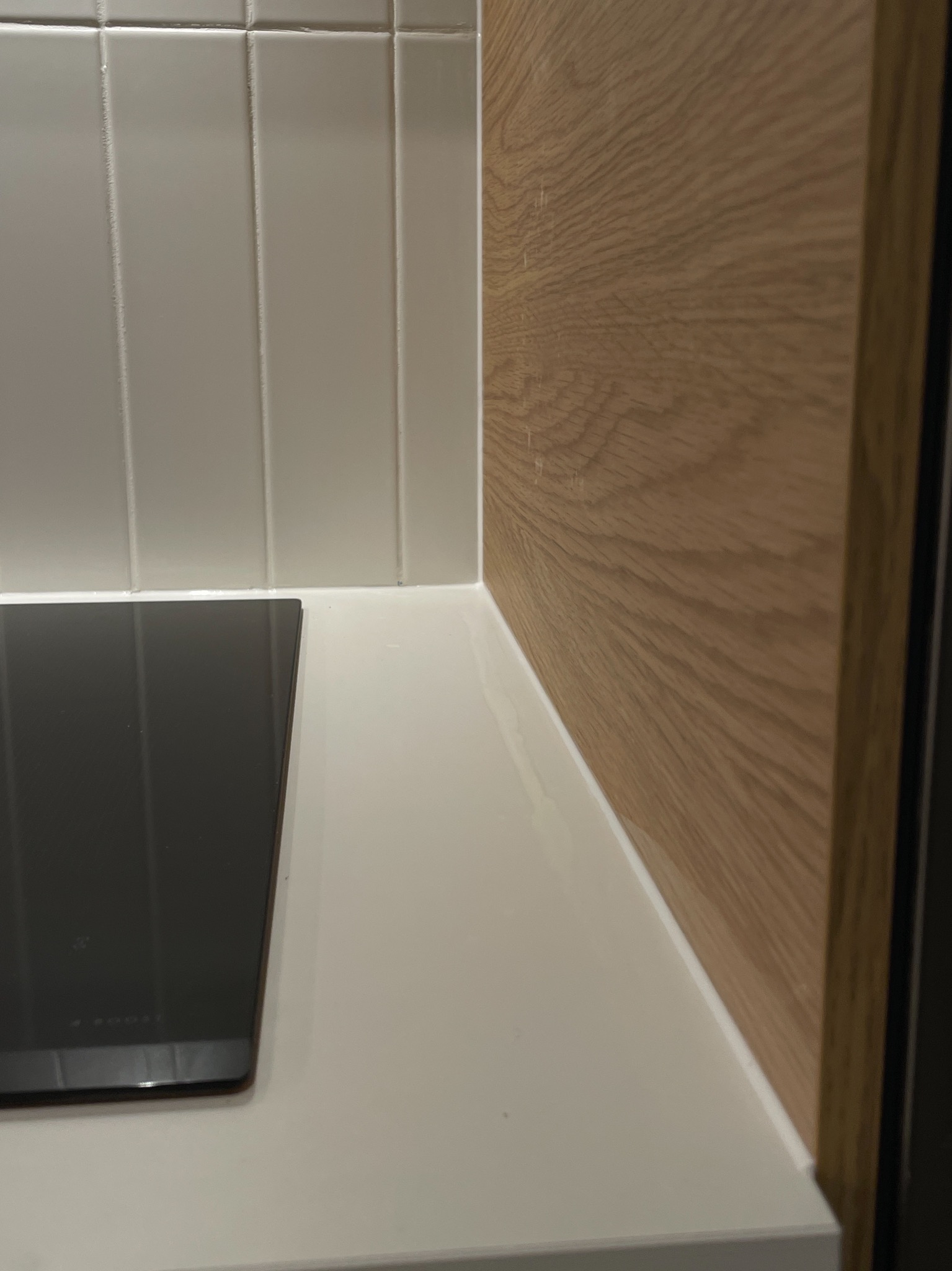 Minimalistyczna kuchnia: białe płytki ścienne, drewniana okładzina i czarna płyta indukcyjna na białym blacie. Nowoczesne wykończenie.