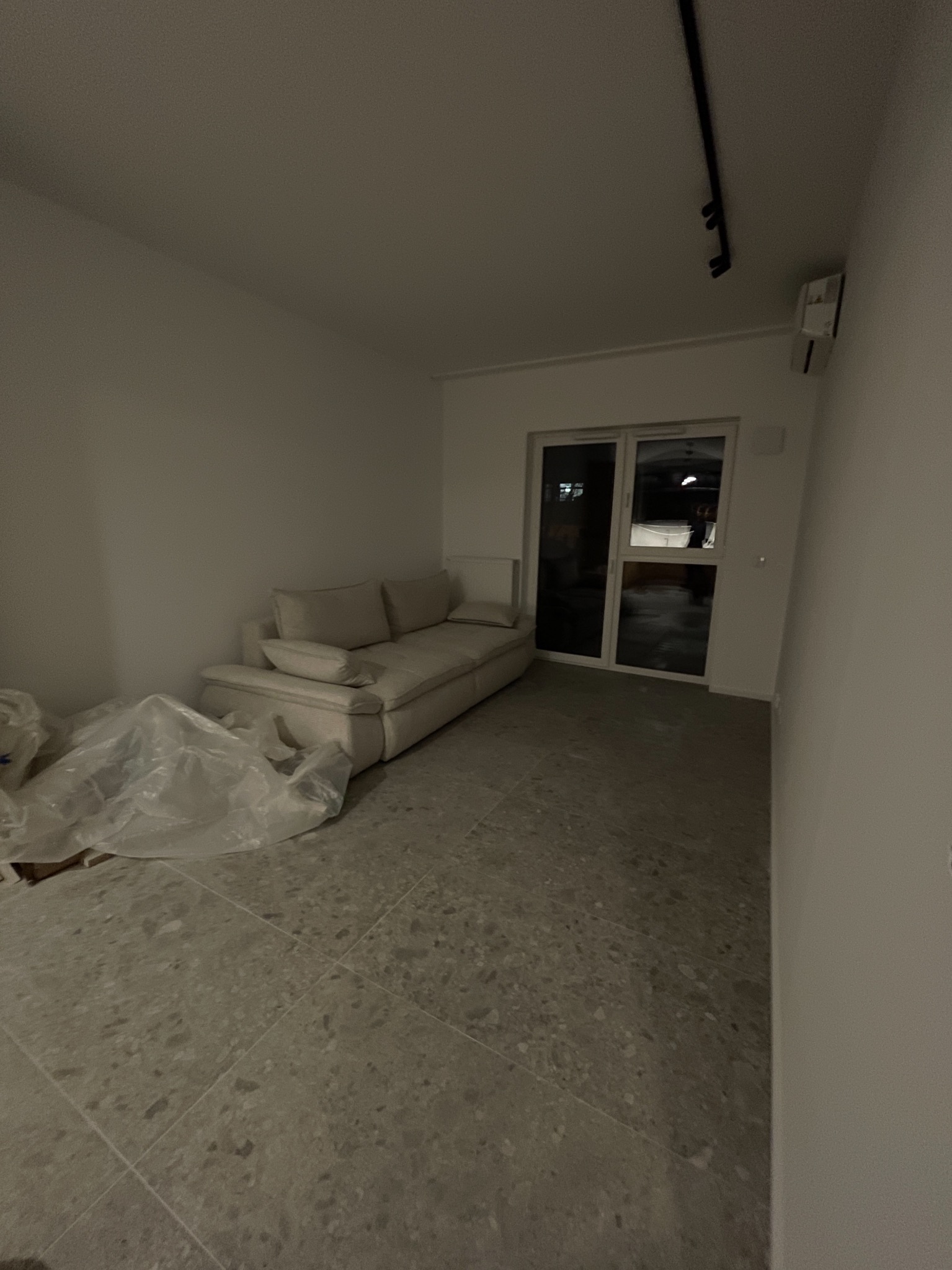 Jasne wnętrze w trakcie wykańczania: szara podłoga z terazzo, sofa pod oknem balkonowym, folia ochronna na podłodze. Minimalistyczne oświetlenie sufitowe.
