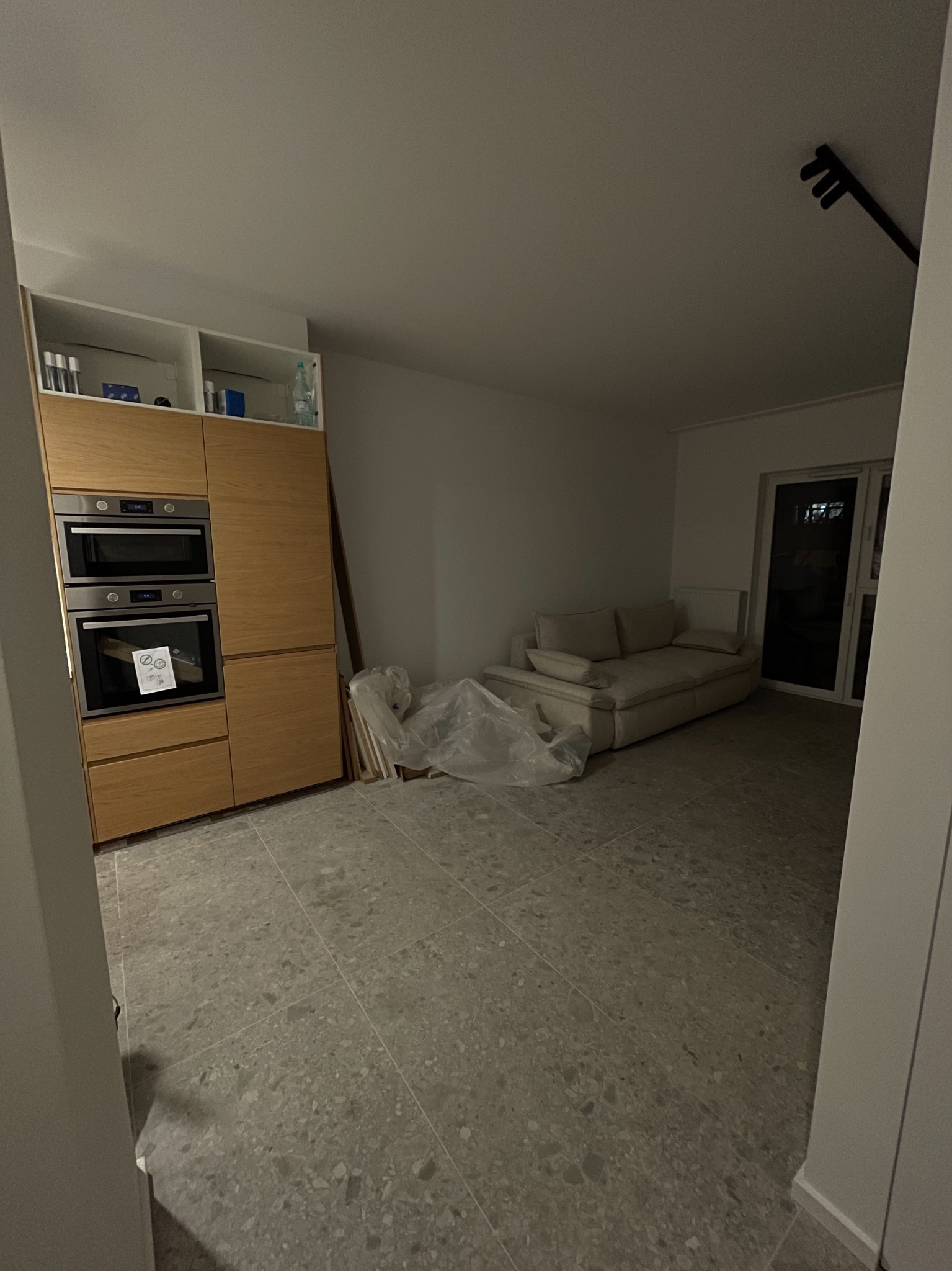 Jasne wnętrze po remoncie: szara podłoga z terazzo, zabudowa kuchenna z drewnianymi frontami, sofa w tle. Część pomieszczenia przykryta folią ochronną.