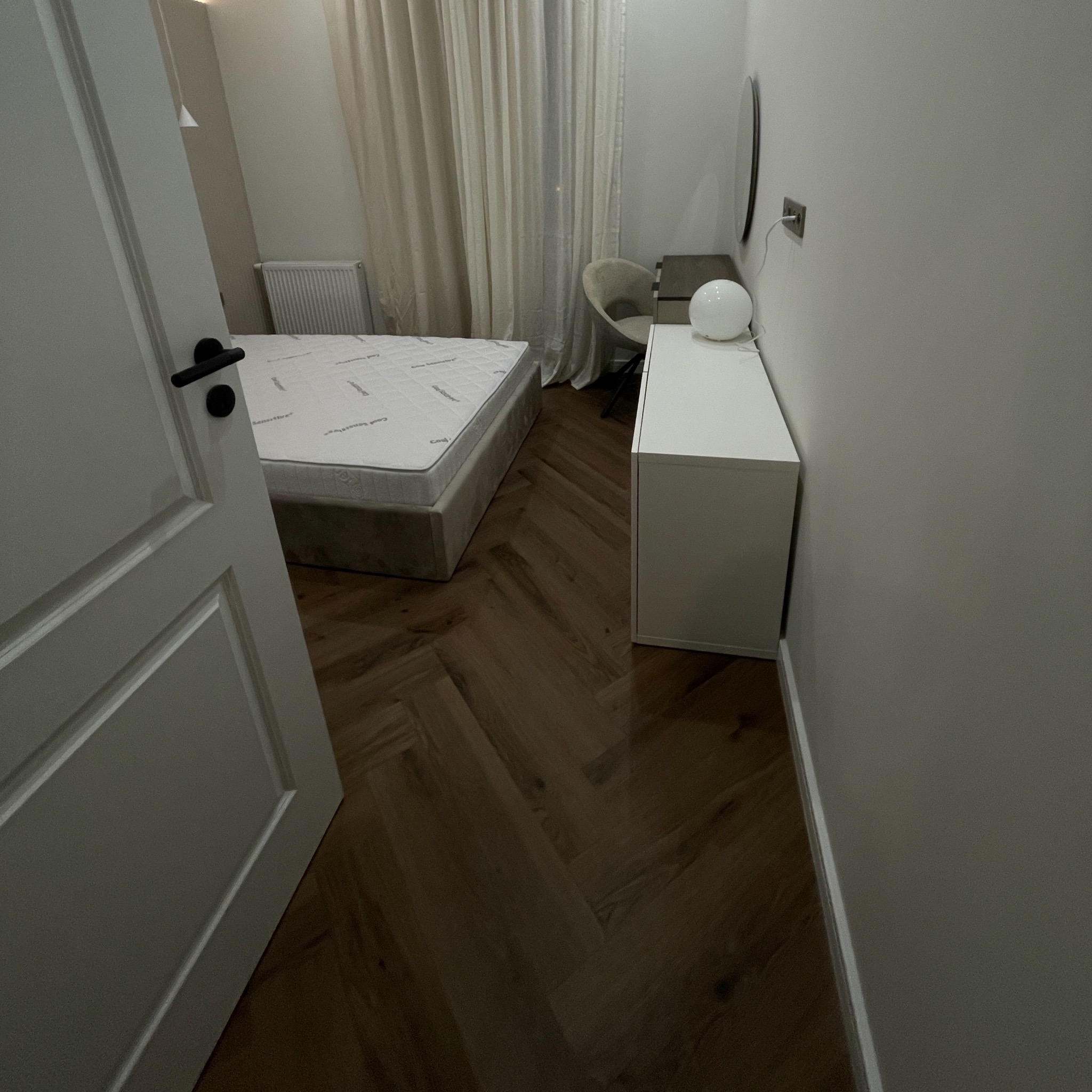 Nowoczesna sypialnia z parkietem ułożonym w jodełkę, widok zza uchylonych drzwi. Neutralna kolorystyka ścian i zasłon, białe meble i łóżko.