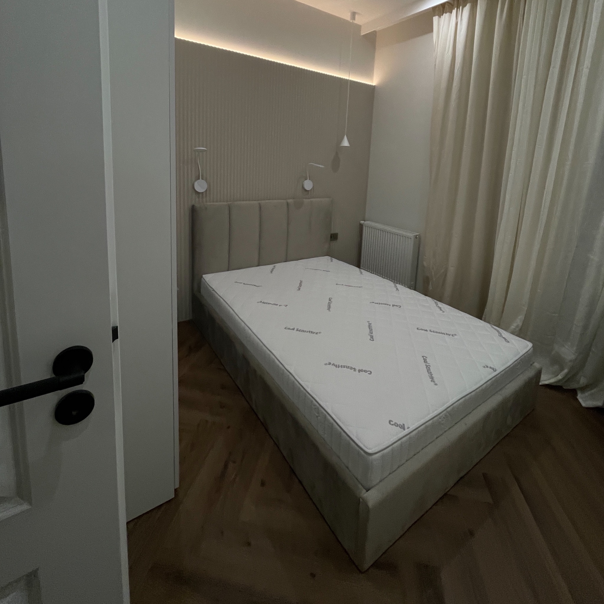 Nowoczesna sypialnia z beżowym łóżkiem, panelami ściennymi i subtelnym oświetleniem LED. Parkiet w jodełkę dodaje elegancji. Widok zza uchylonych drzwi.