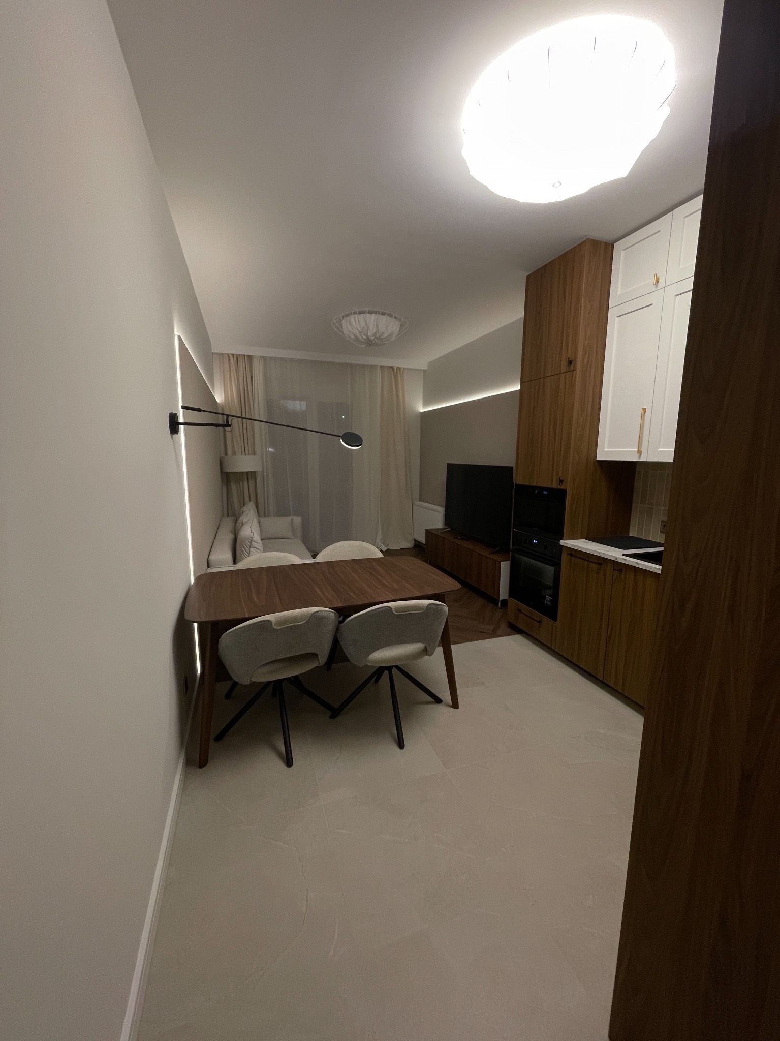 Nowoczesne wnętrze: stół z krzesłami, zabudowa kuchenna z drewnianymi frontami i białymi szafkami, oświetlenie LED wzdłuż ściany, jasna podłoga i sofa w tle.