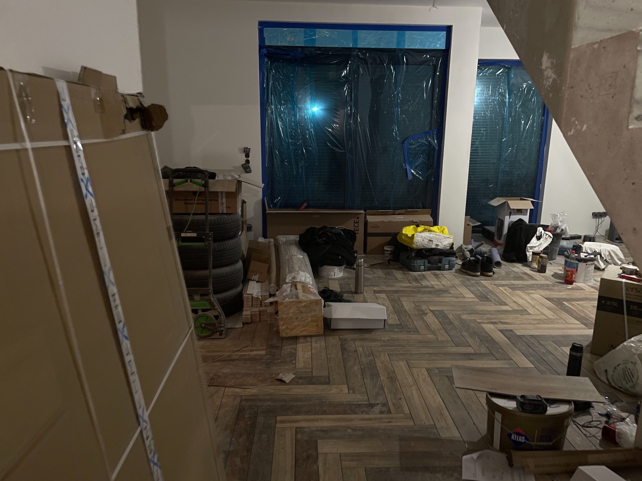 Wnętrze w trakcie remontu: zabezpieczone okna folią, podłoga w jodełkę, materiały budowlane i narzędzia. Widoczne kartony i akcesoria malarskie.