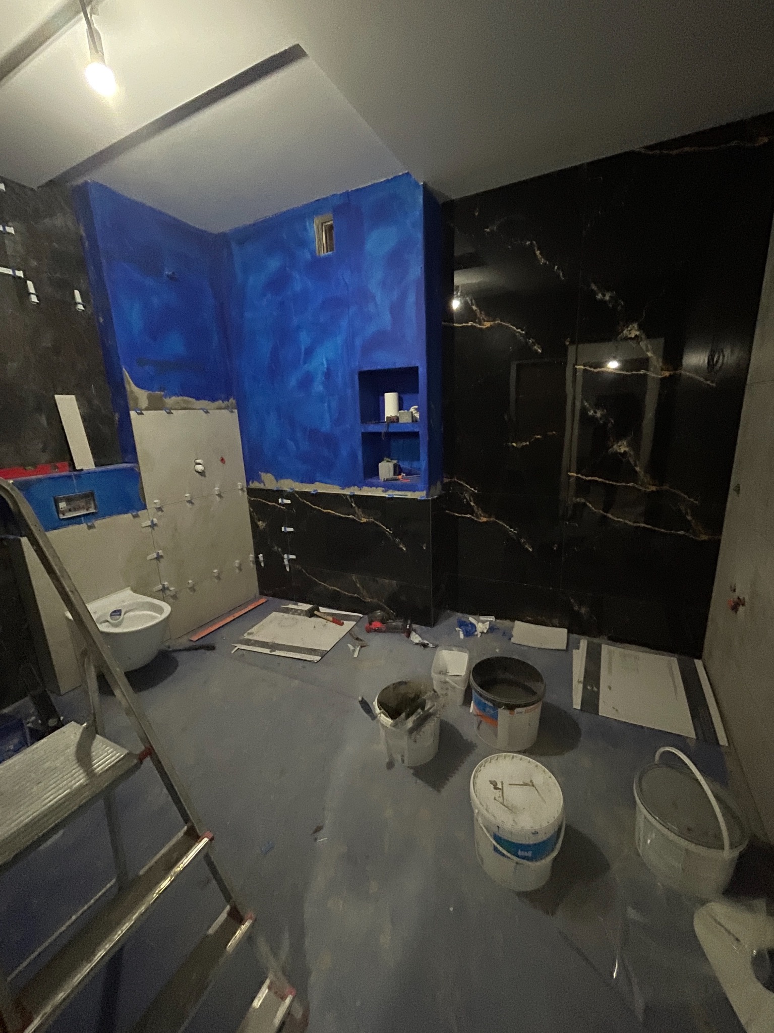 Remont łazienki w trakcie: częściowo położone czarne płytki imitujące marmur, niebieska ściana z wnęką, widoczne narzędzia i drabina.