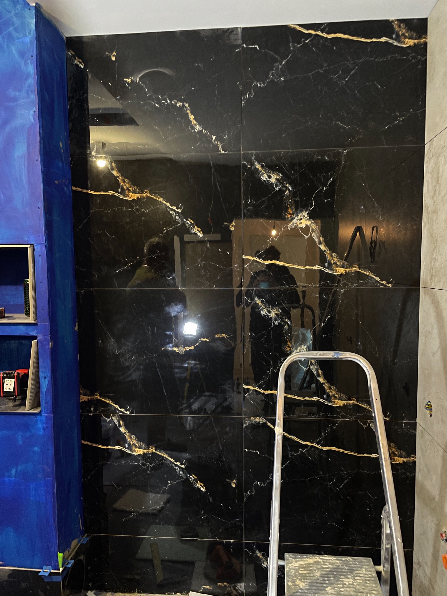 Ściana wyłożona czarnymi, błyszczącymi płytkami z złotymi żyłami, częściowo widoczna drabina i niebieska obudowa. Prace w toku, widoczne elementy montażowe.