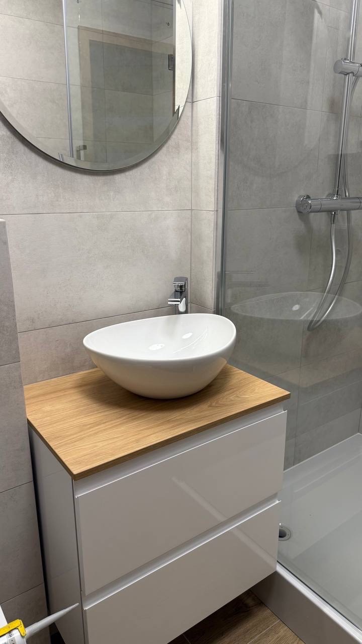 Nowoczesna łazienka: owalna umywalka nablatowa na szafce z drewnianym blatem, szare płytki, okrągłe lustro i kabina prysznicowa w tle.