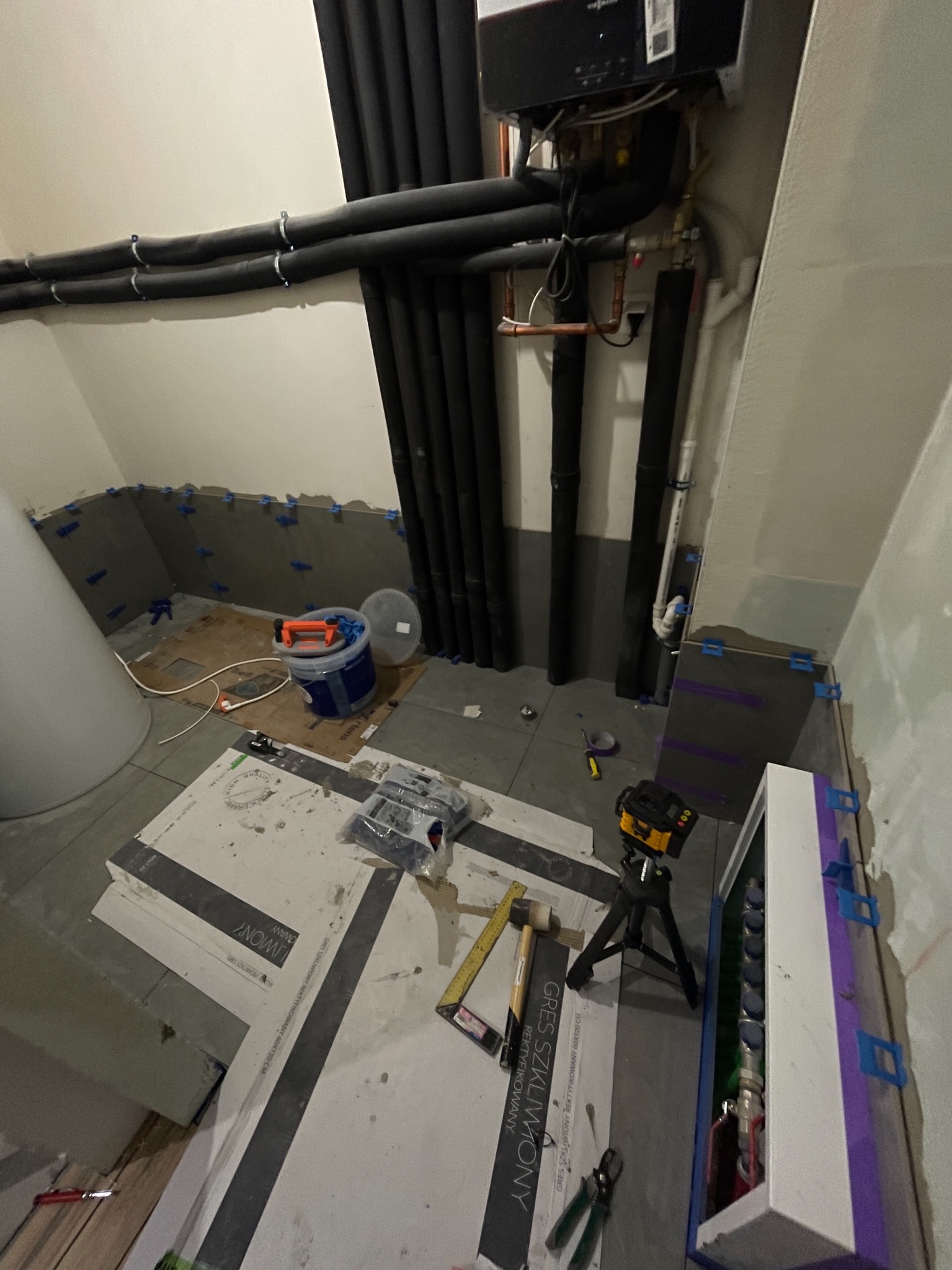 Remont łazienki w trakcie: ułożone płytki podłogowe i ścienne z użyciem klipsów poziomujących, widoczne rury instalacji, narzędzia i laser krzyżowy na statywie.