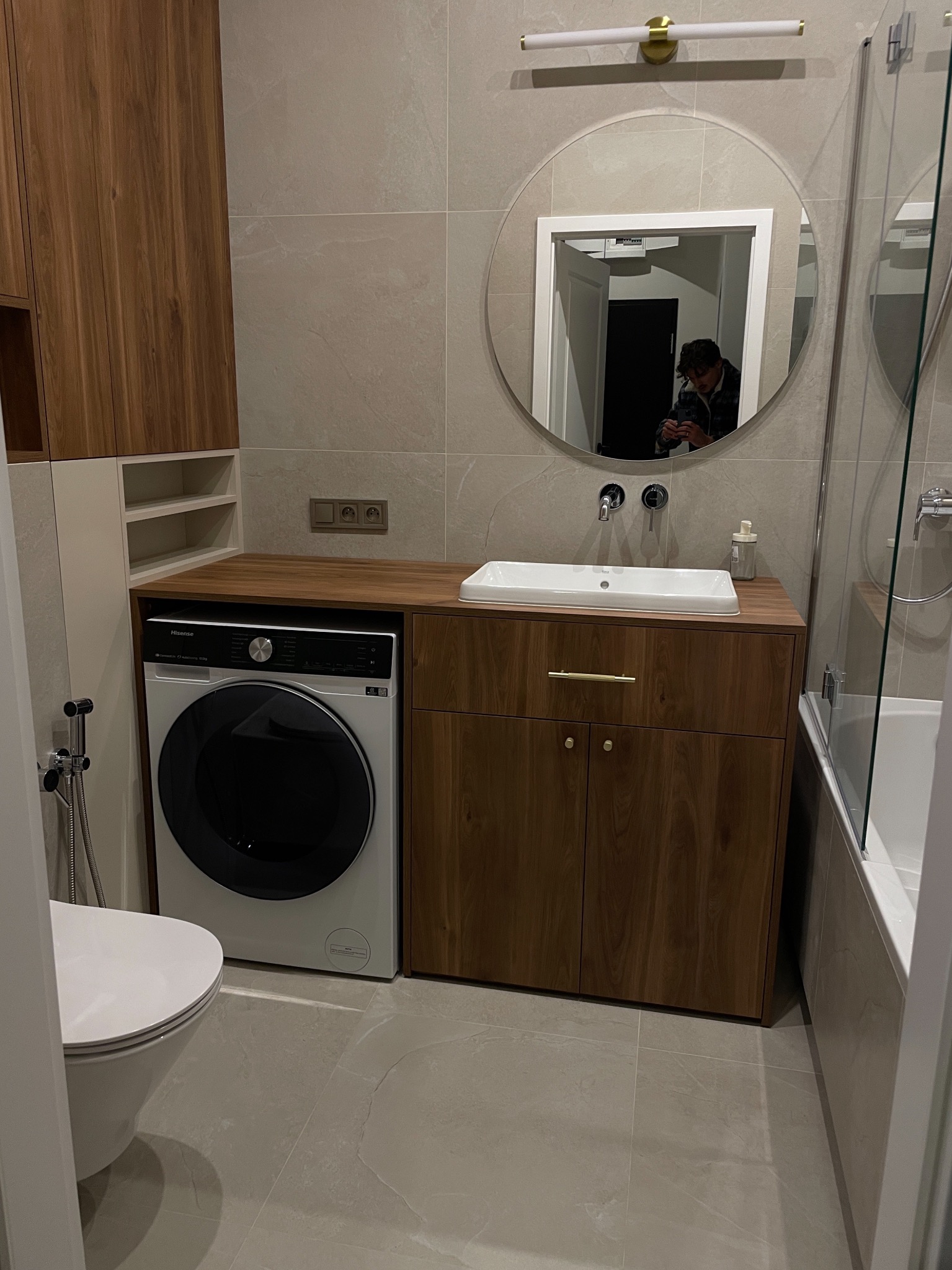 Nowoczesna łazienka z drewnianymi szafkami, okrągłym lustrem z podświetleniem i pralką w zabudowie. Jasne płytki i minimalistyczny design.