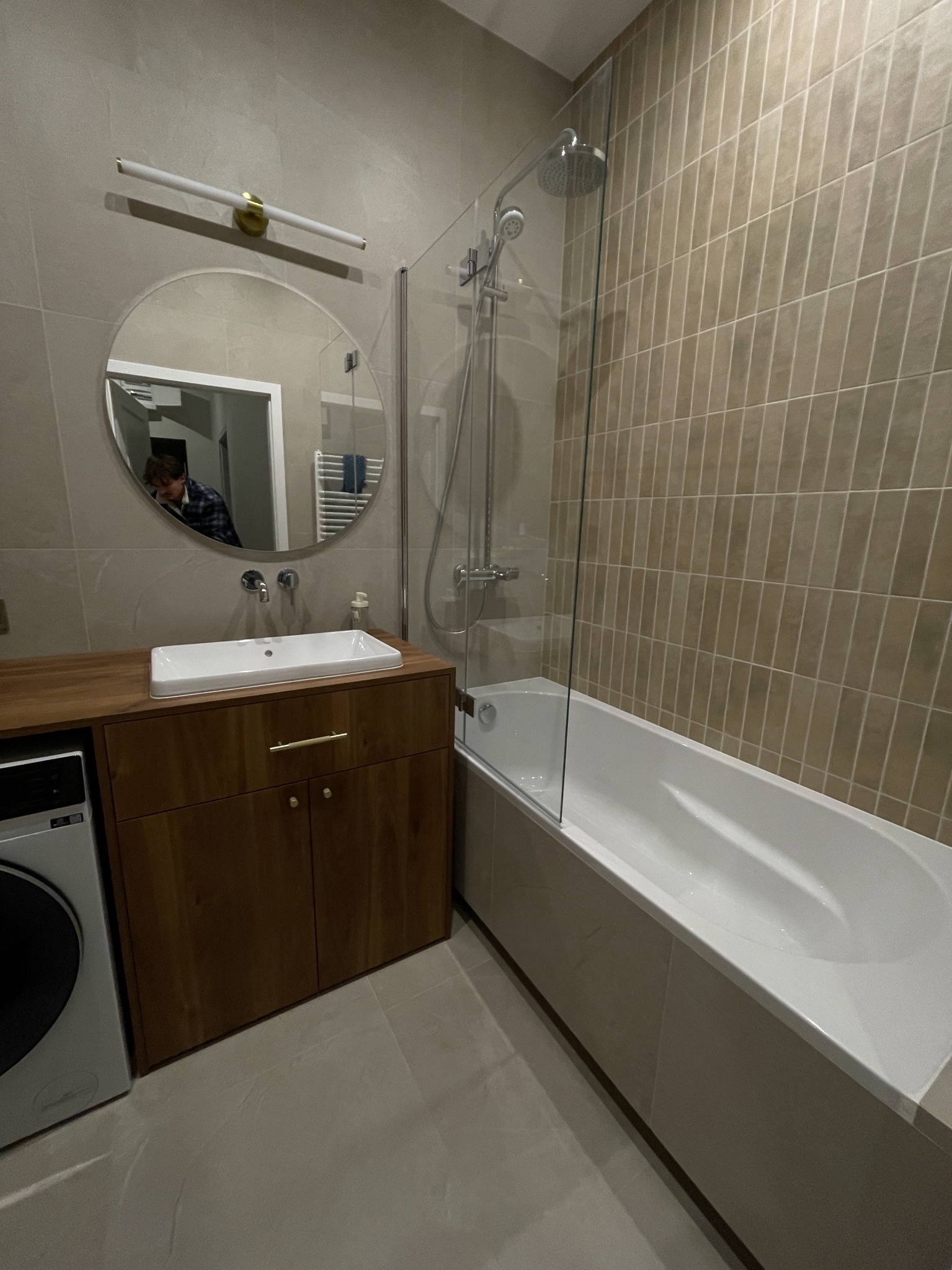 Nowoczesna łazienka z beżowymi płytkami, białą wanną z parawanem, drewnianą szafką pod umywalką i okrągłym lustrem z oświetleniem LED.