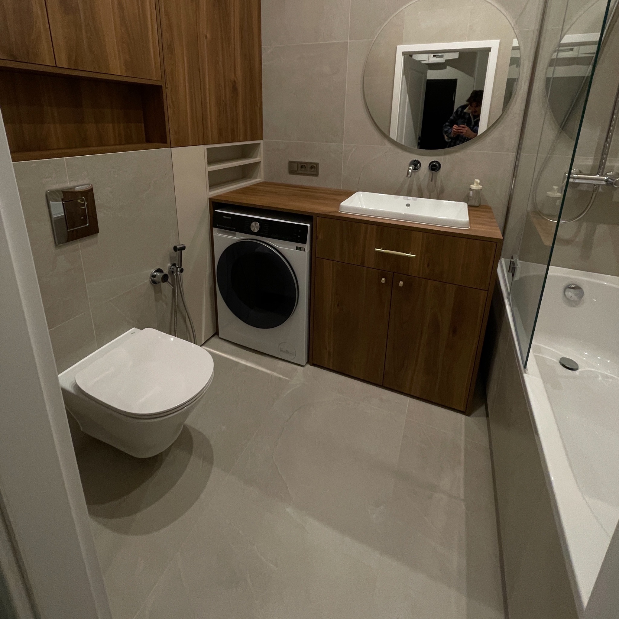Nowoczesna łazienka z drewnianymi akcentami: umywalka z szafką, pralka pod blatem, wisząca toaleta i okrągłe lustro. Całość utrzymana w jasnej, stonowanej kolorystyce.