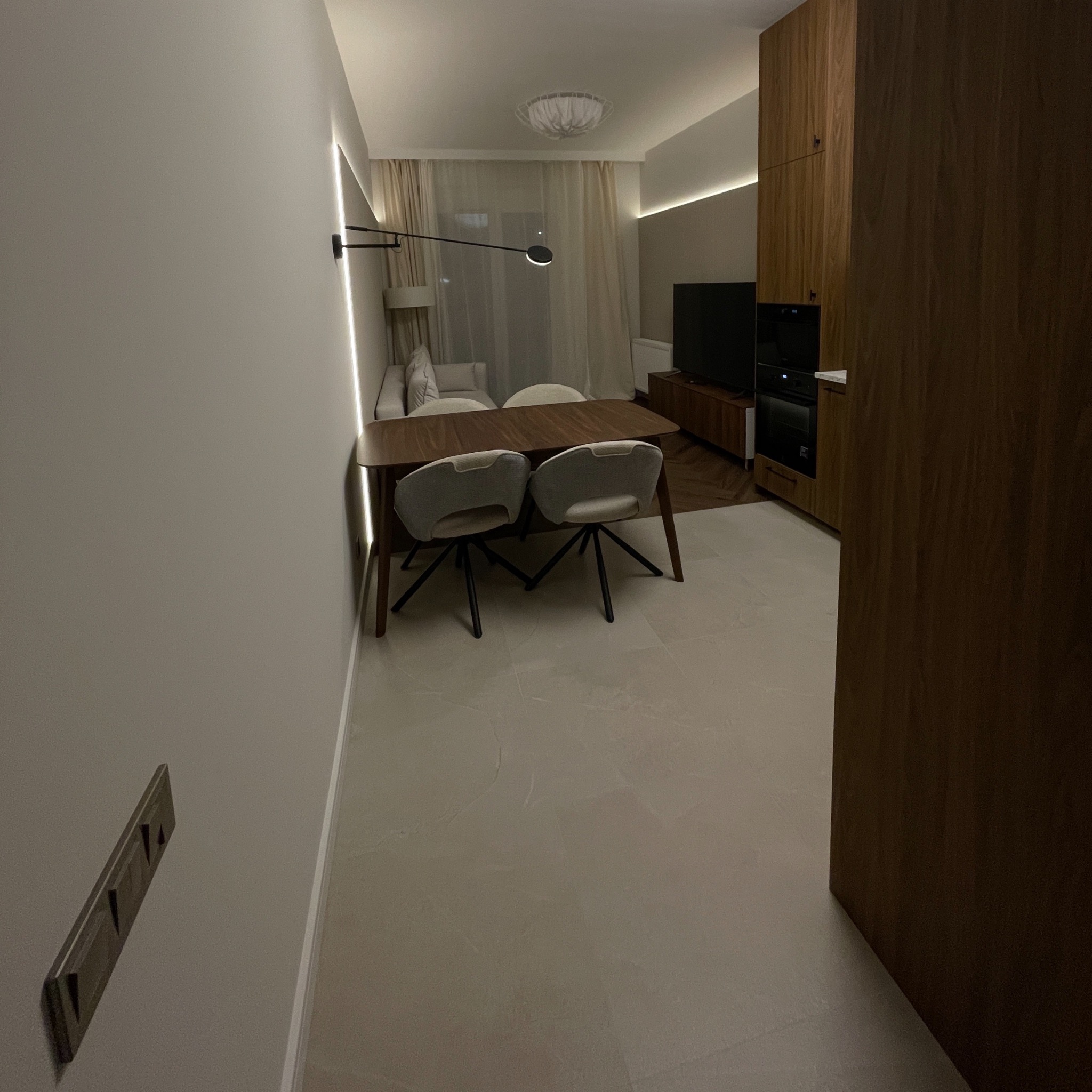 Nowoczesne wnętrze mieszkania z drewnianym stołem i krzesłami, oświetlone listwami LED. Widoczna zabudowa kuchenna i elementy dekoracyjne.