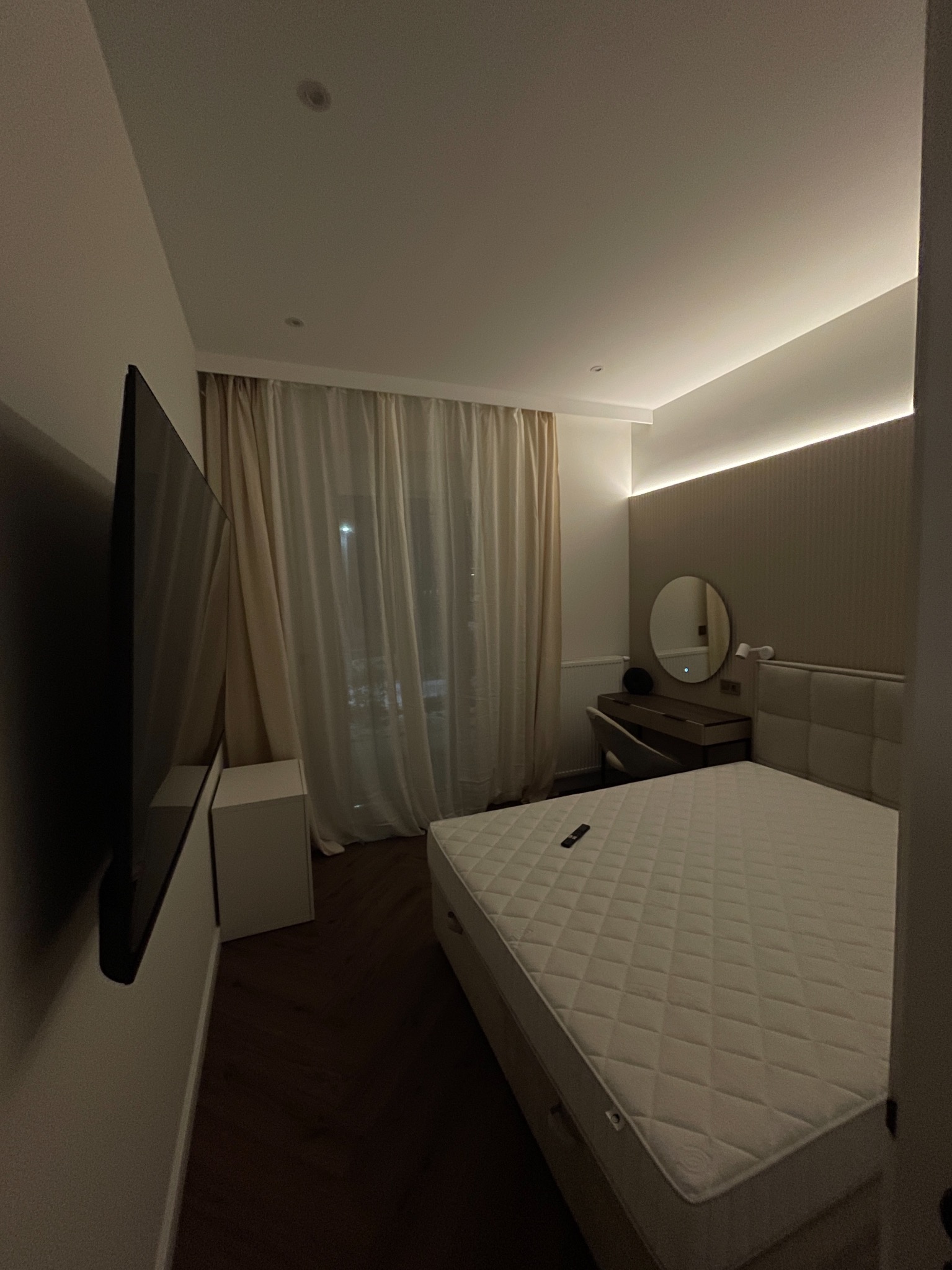 Nowoczesna sypialnia z dużym telewizorem na ścianie, łóżkiem z pikowanym zagłówkiem i toaletką z okrągłym lustrem, oświetlona listwą LED.