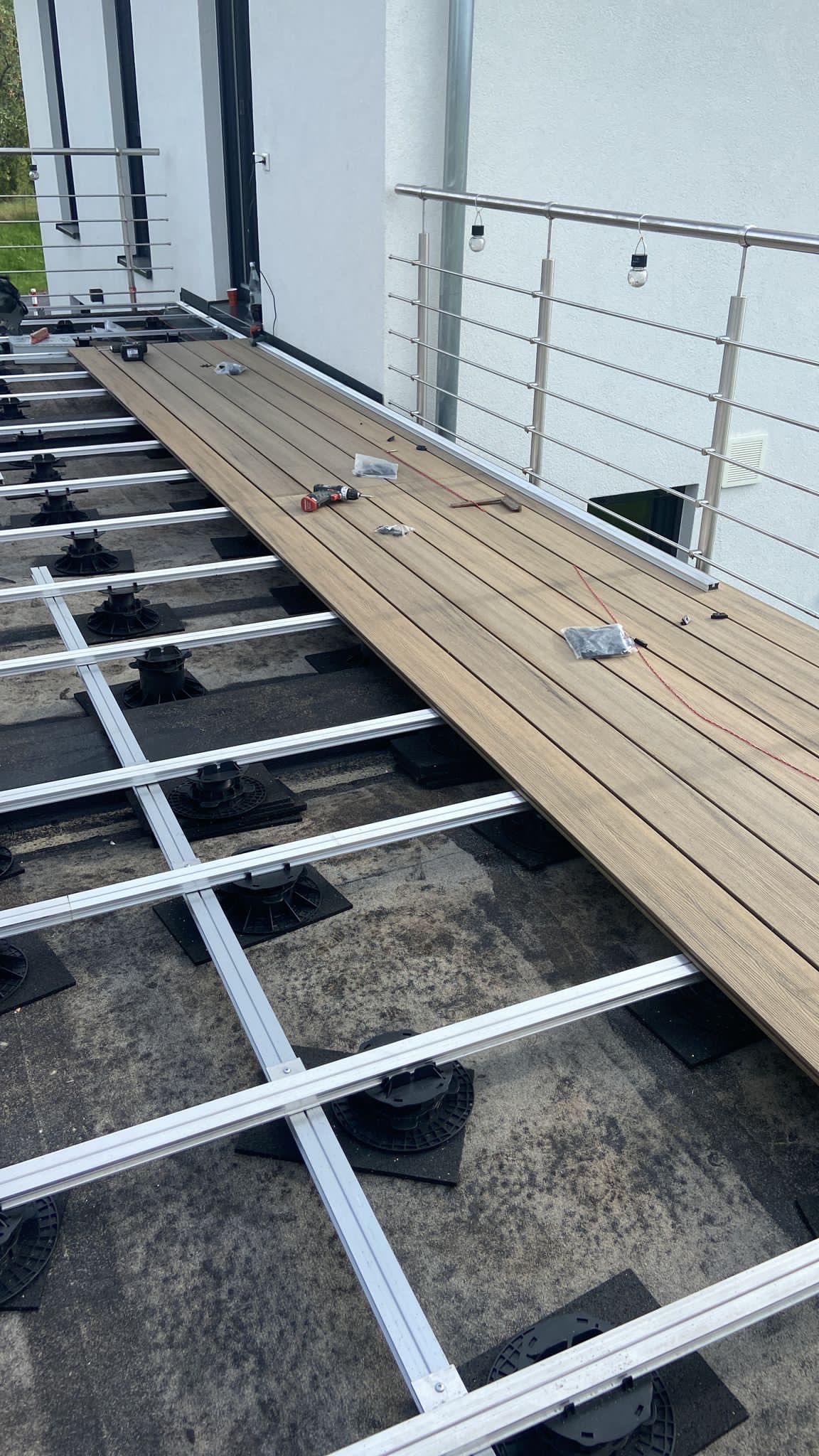 Montaż desek kompozytowych na regulowanych wspornikach z aluminiową podkonstrukcją na balkonie, widoczne narzędzia i akcesoria montażowe.