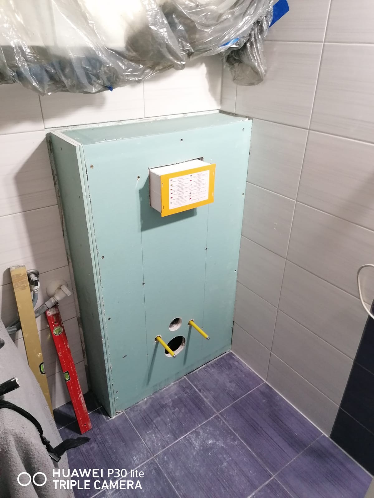 Konstrukcja z płyty kartonowo-gipsowej zabudowująca stelaż WC w trakcie montażu, widoczne otwory na przyłącza, poziomica i folia ochronna na ścianie.