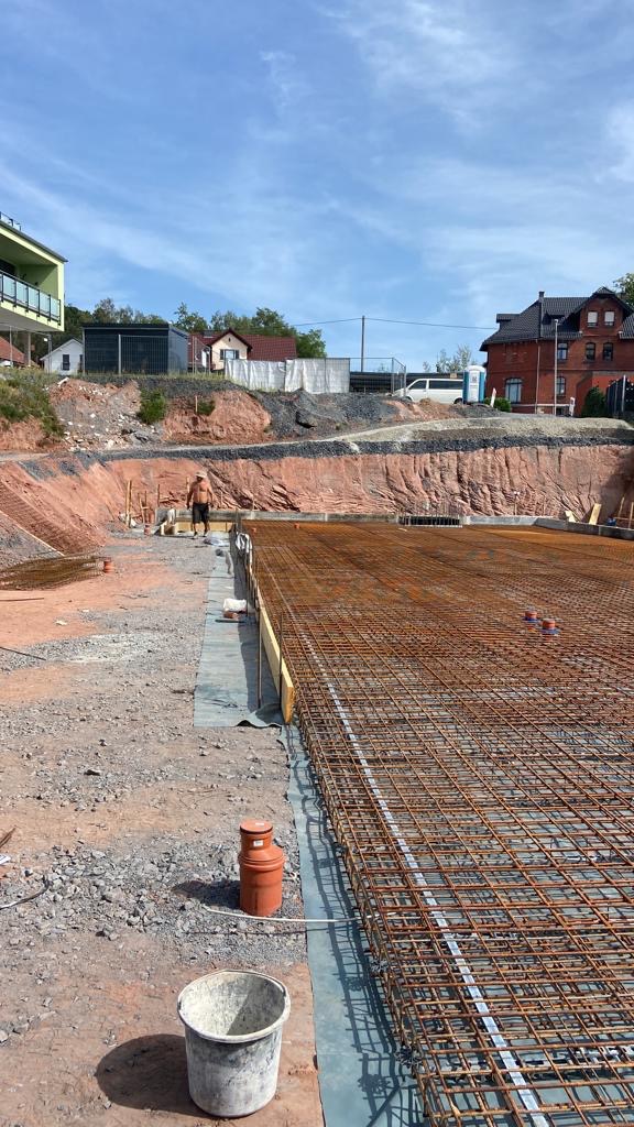 Zbrojenie fundamentów pod budynek, widoczne stalowe pręty tworzące siatkę, szalunki z desek, pomarańczowa butla, wiadro i pracownik w tle na tle wykopu i zabudowy mieszkalnej.
