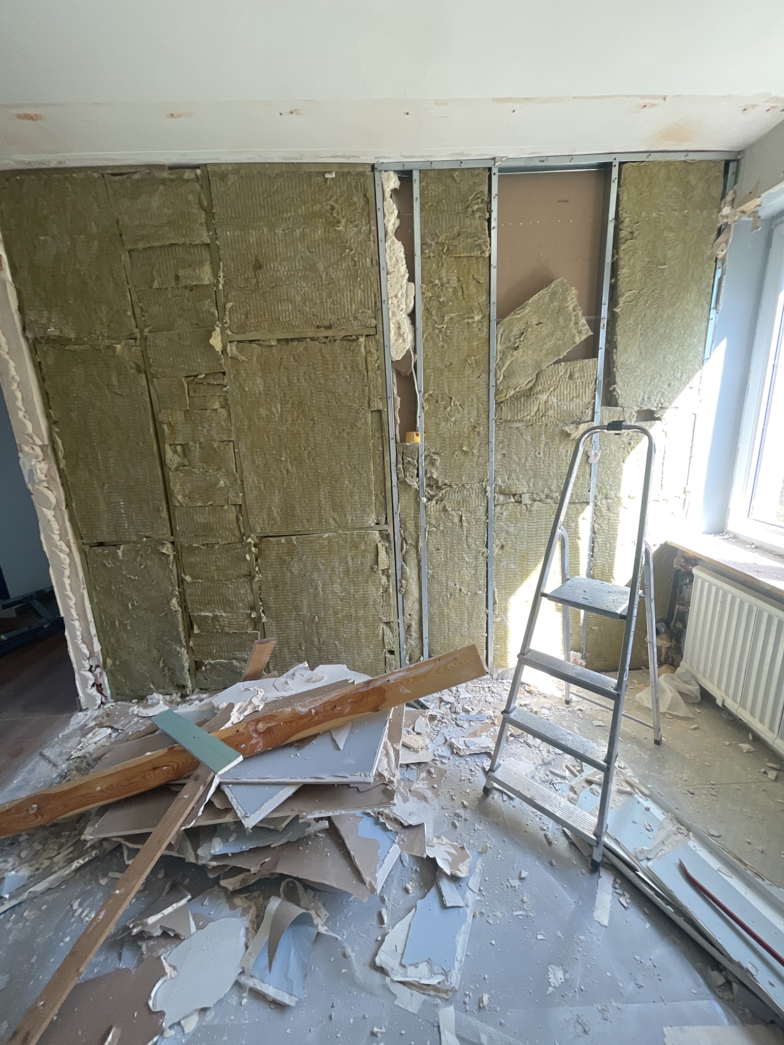 Montaż izolacji z wełny mineralnej w metalowym stelażu ściany, widoczne fragmenty starej okładziny i drabina aluminiowa.