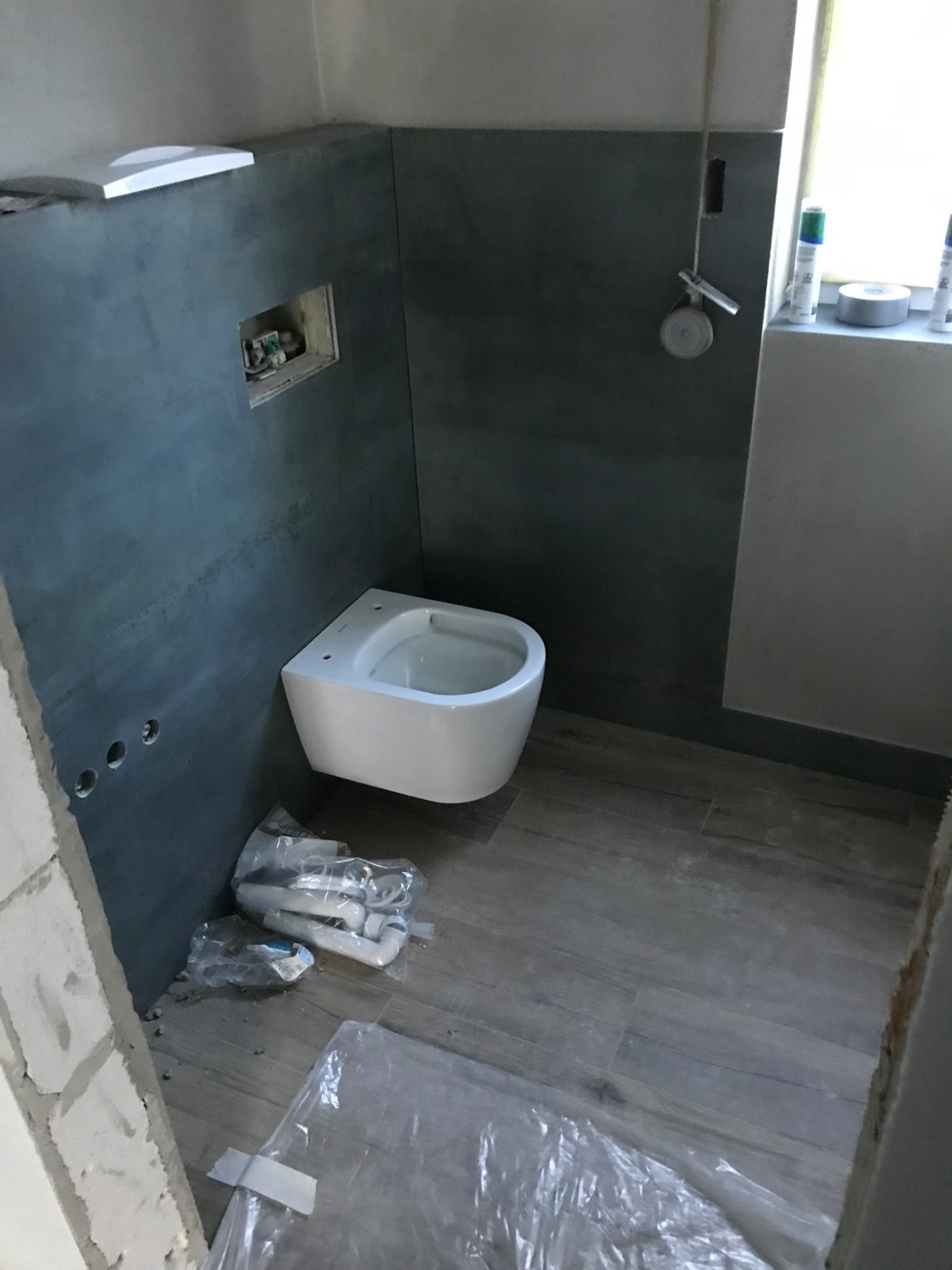 Montaż podwieszanej toalety na tle szarych płytek w trakcie remontu łazienki, widoczne rury i elementy instalacji w folii ochronnej, oraz częściowo wykończona ściana z pustaków.