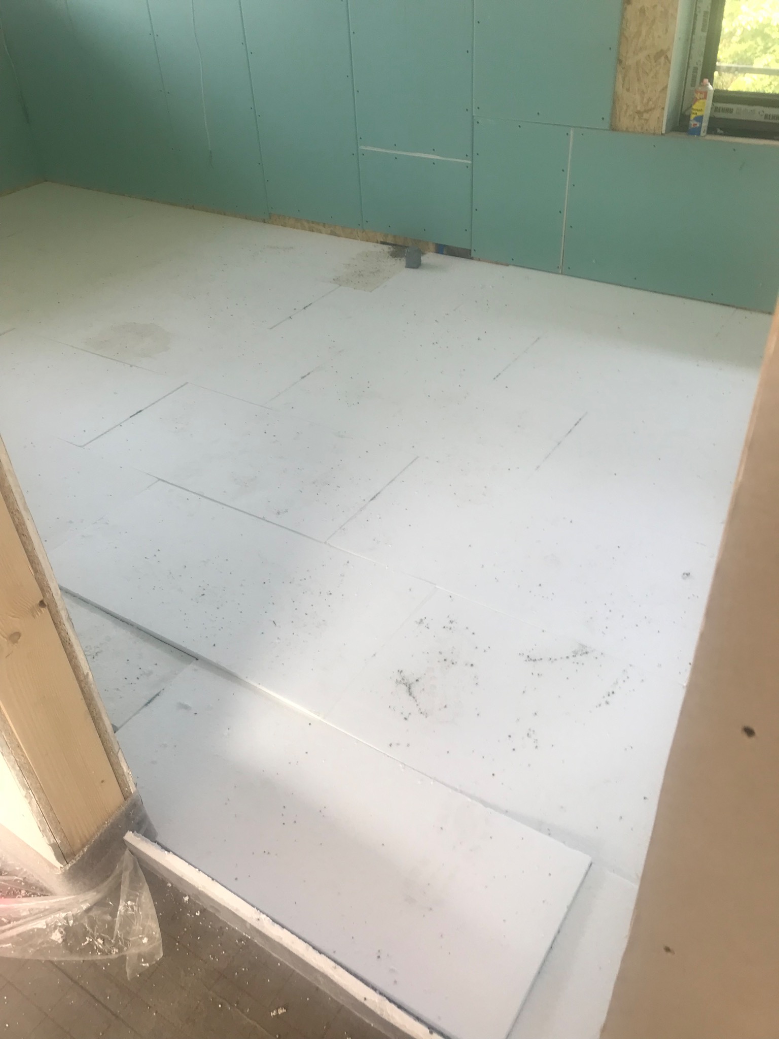 Pokój w trakcie prac wykończeniowych: ściany pokryte zielonymi płytami, podłoga wyłożona białymi płytami styropianowymi, widoczne okno z preparatami na parapecie.