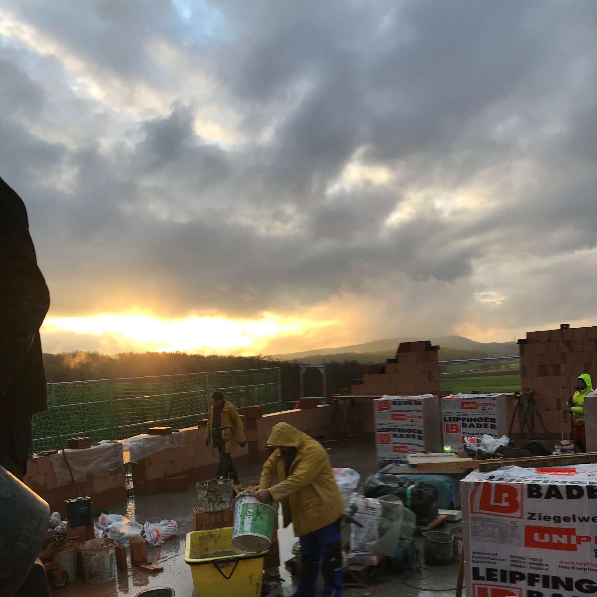 Budowa domu w trakcie, murarze w żółtych kurtkach roboczych przygotowują zaprawę, w tle cegły i zachmurzone niebo z przebijającym się słońcem.