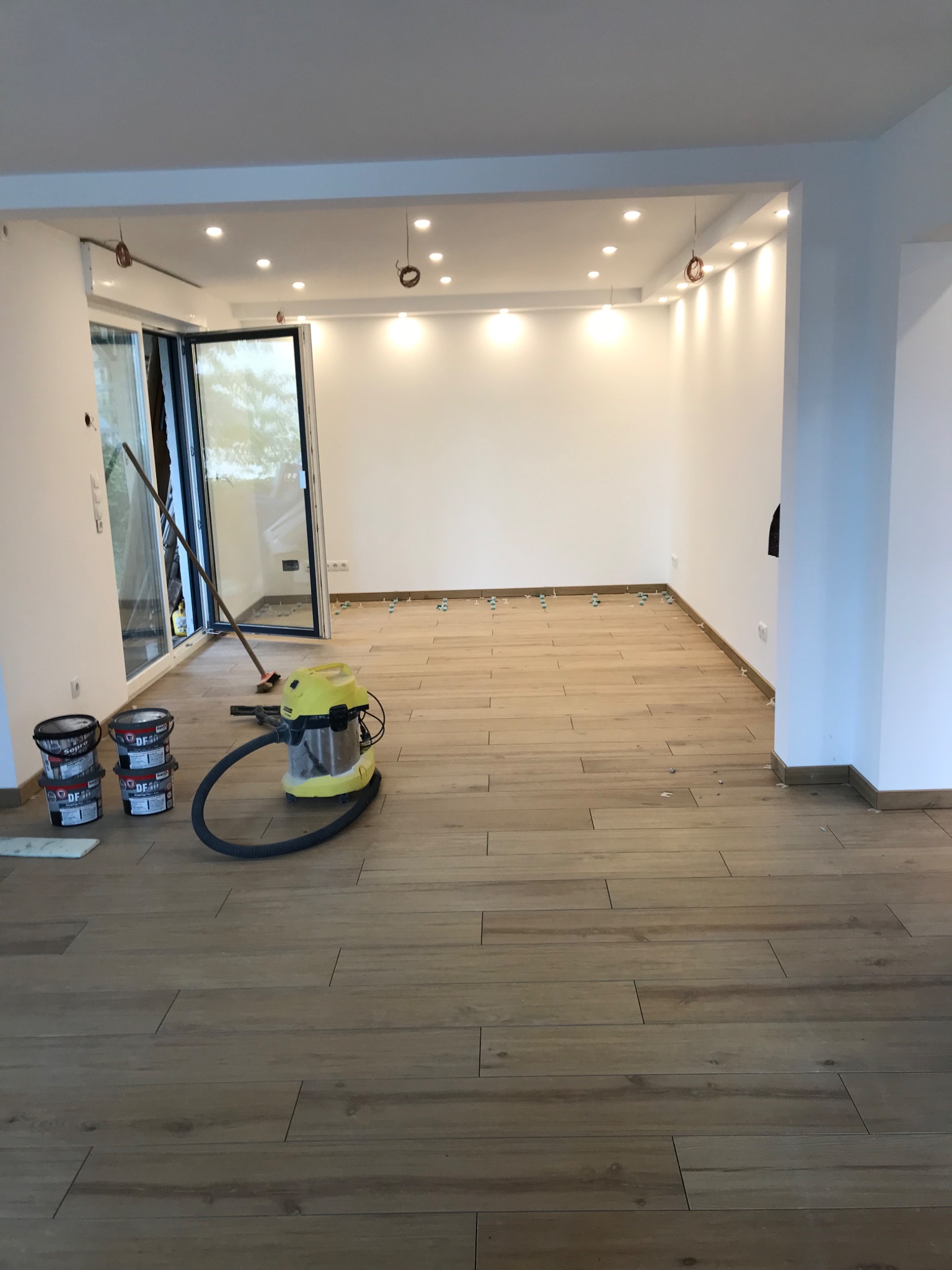 Jasne, drewnopodobne płytki podłogowe ułożone w nowym pomieszczeniu, widoczne pojemniki z klejem, odkurzacz przemysłowy oraz krzyżyki dystansowe, otwarte okno balkonowe.