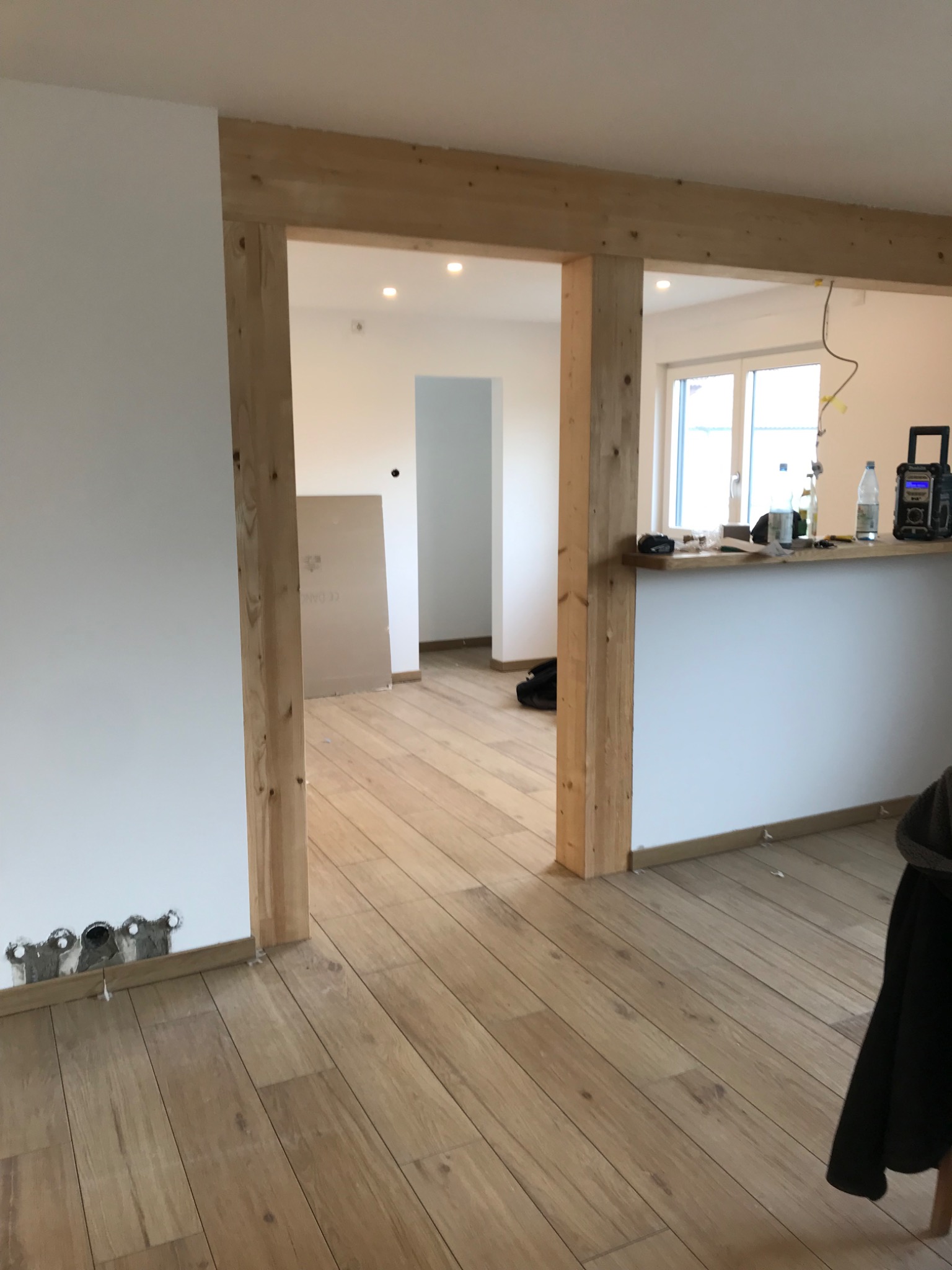 Wnętrze w trakcie prac wykończeniowych: drewniana konstrukcja ramy drzwiowej, świeżo położona podłoga z paneli, przygotowane otwory na gniazdka elektryczne w ścianie, płyta gipsowa oparta o ścianę...