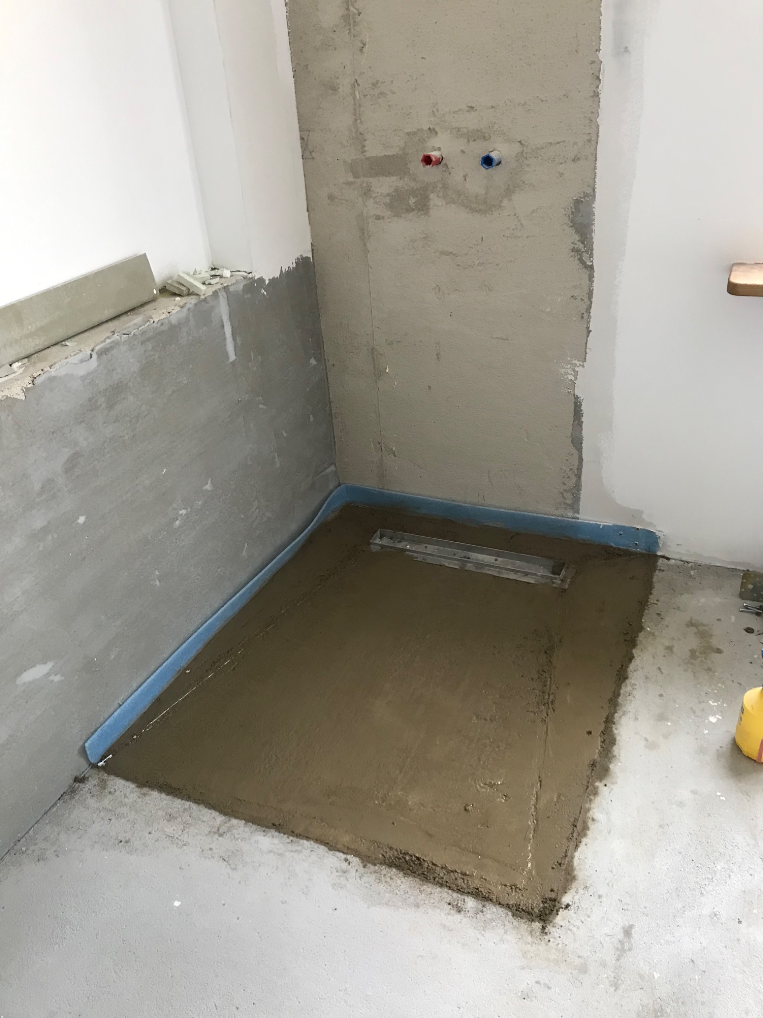 Przygotowanie podłoża pod prysznic z odpływem liniowym, z widoczną hydroizolacją w narożnikach, surowy beton na ścianach i częściowo pomalowana ściana obok.