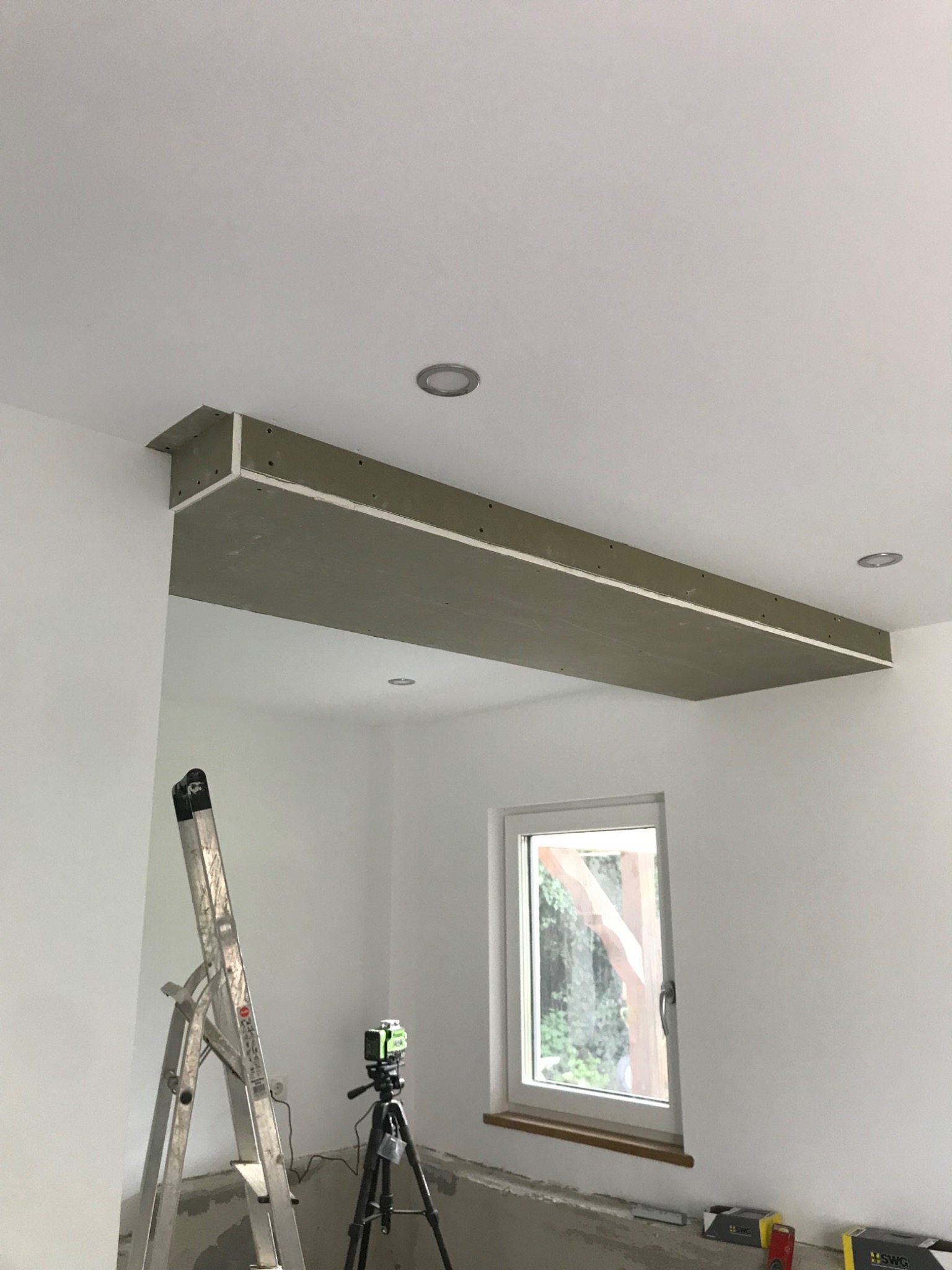 Montaż podwieszanej konstrukcji sufitowej z płyt gipsowo-kartonowych, widoczna drabina, laser krzyżowy na statywie i fragment okna
