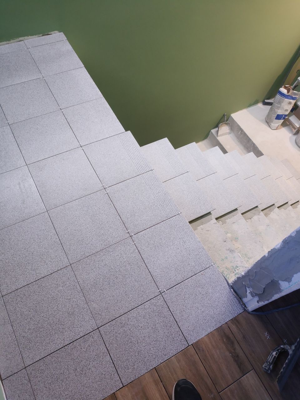 Schody w trakcie układania szarych, kwadratowych płytek, widok z góry, z zieloną ścianą w tle i workiem zaprawy w rogu, częściowo wykończone drewnem na dole.