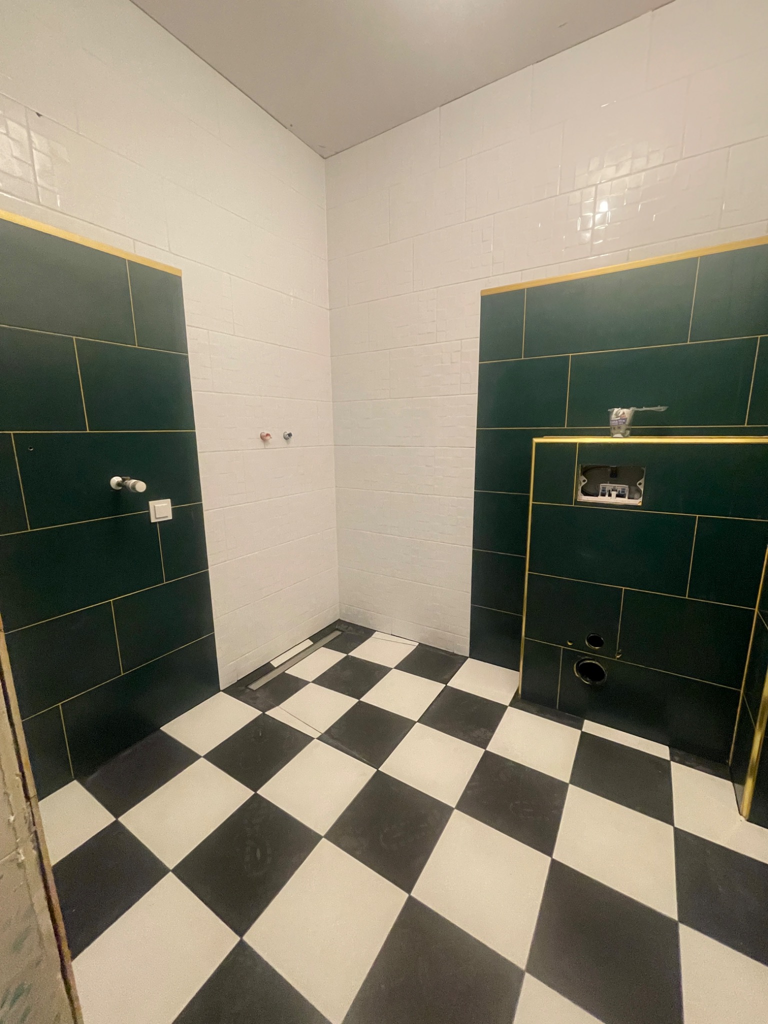 Wnętrze łazienki w trakcie prac wykończeniowych: zielone i białe płytki ścienne z złotymi listwami, czarno-biała szachownica na podłodze, widoczne instalacje wodno-kanalizacyjne.