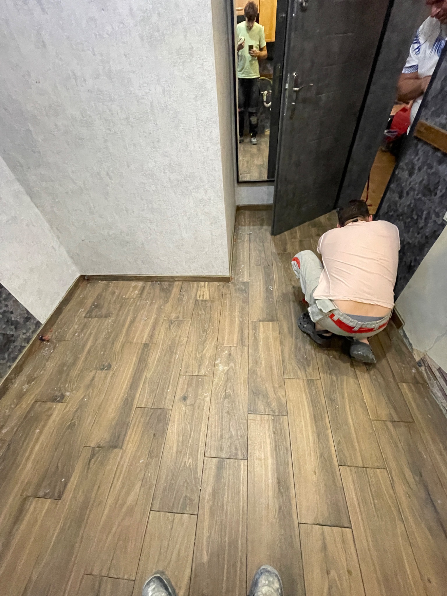 Układanie podłogi z płytek imitujących drewno w korytarzu, widoczne niedociągnięcia przy ścianie, osoba w trakcie pracy, odbicie w lustrze.