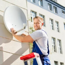 Uśmiechnięty monter w niebieskim kombinezonie, stojący na czerwonej drabinie, przytwierdza białą antenę satelitarną do elewacji budynku.