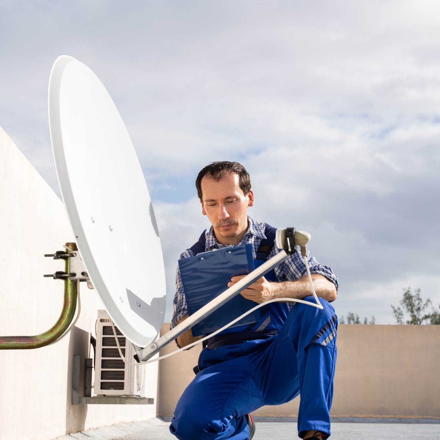 Instalator w niebieskim kombinezonie klęczy obok zamontowanej anteny satelitarnej, trzymając niebieski clipboard. W tle widoczny klimatyzator na ścianie i pochmurne niebo.