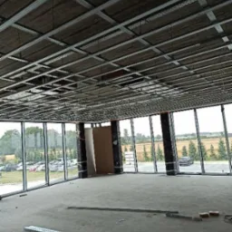 Montaż metalowej konstrukcji sufitu podwieszanego w przestronnym pomieszczeniu z dużymi oknami, widok na zewnątrz z zielenią i parkingiem, elementy konstrukcyjne leżące na surowej podłodze.