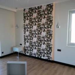 Pokój po remoncie z tapetą w kwiaty na jednej ze ścian, drewnianą podłogą, nowoczesnym oświetleniem i niedokończonym meblem na podłodze.