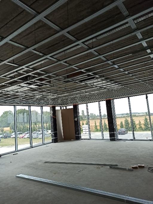 Montaż metalowej konstrukcji sufitu podwieszanego w przestronnym pomieszczeniu z dużymi oknami, widok na zewnątrz z zielenią i parkingiem, elementy konstrukcyjne leżące na surowej podłodze.