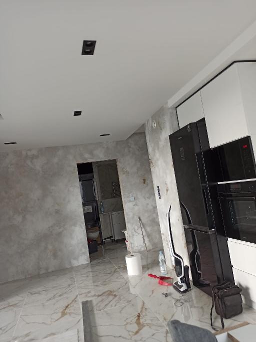 Wnętrze mieszkania w trakcie wykańczania: marmurowe płytki podłogowe, ściany z efektem dekoracyjnym, biała zabudowa kuchenna z czarnymi akcentami, widoczne narzędzia i materiały budowlane.
