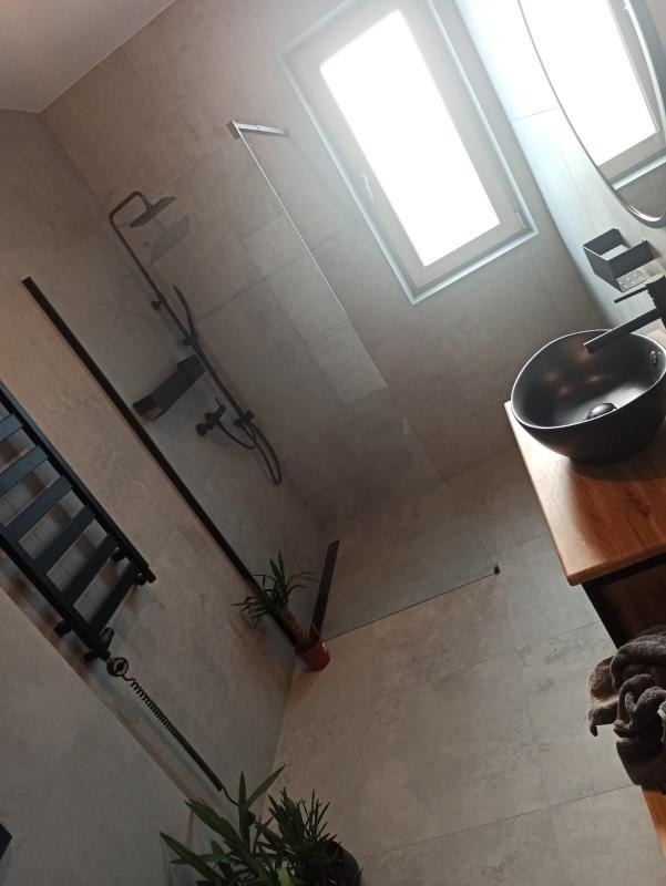 Nowoczesna łazienka z szarą płytką, czarną armaturą i szklanym panelem prysznicowym, uzupełniona roślinami doniczkowymi i drewnianą umywalką.