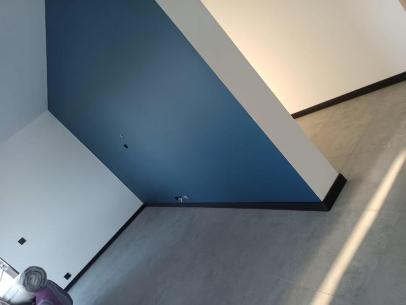 Wnętrze po malowaniu: ściana w kolorze głębokiego błękitu kontrastuje z bielą, wykończona czarnymi listwami przypodłogowymi i szarą podłogą z płytek. Widoczne gniazdka elektryczne w ścianie.