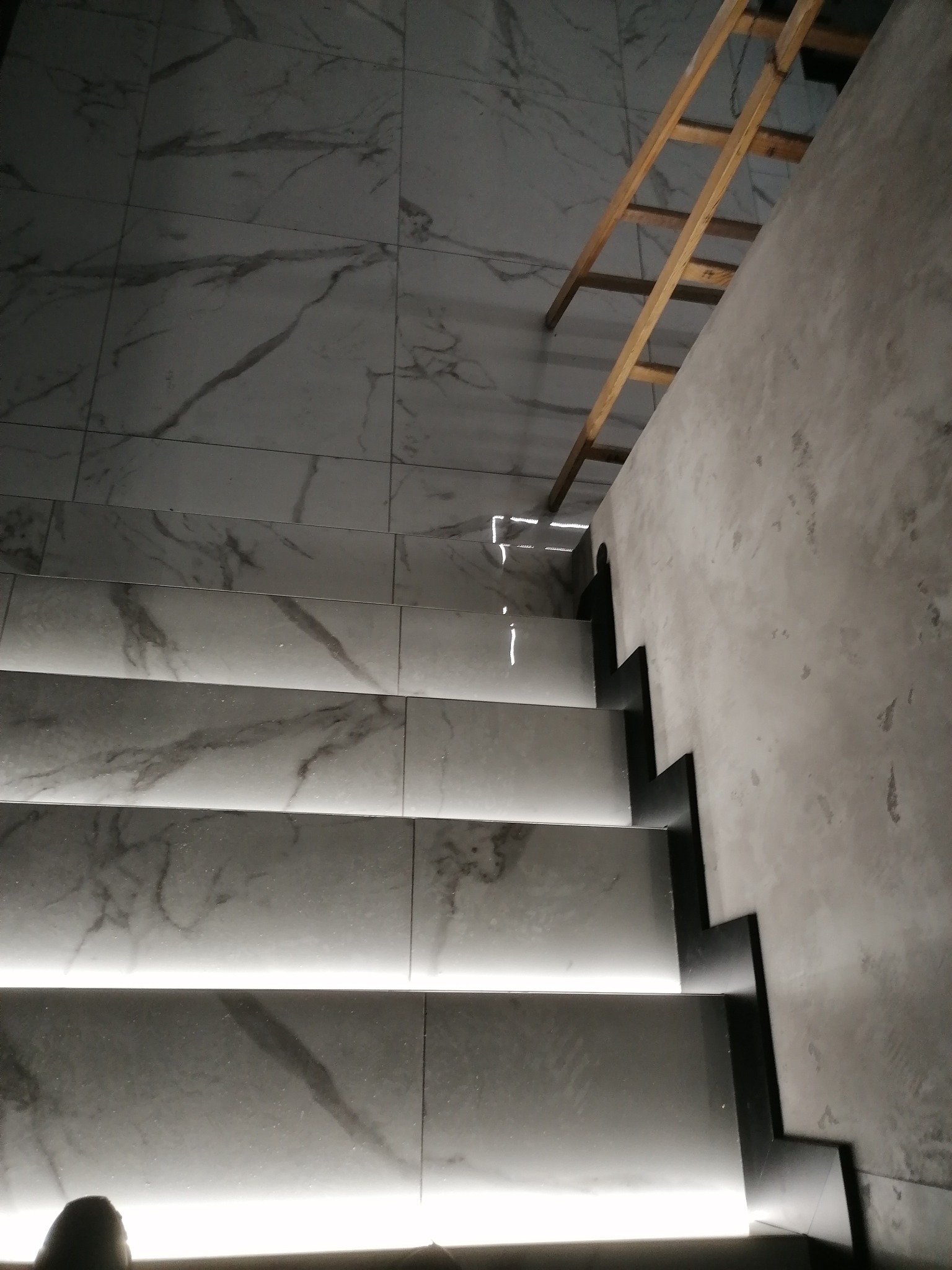 Nowoczesne schody z podświetleniem LED, wykończone płytkami imitującymi marmur, z widoczną drewnianą drabiną w tle i surową, szpachlowaną ścianą.