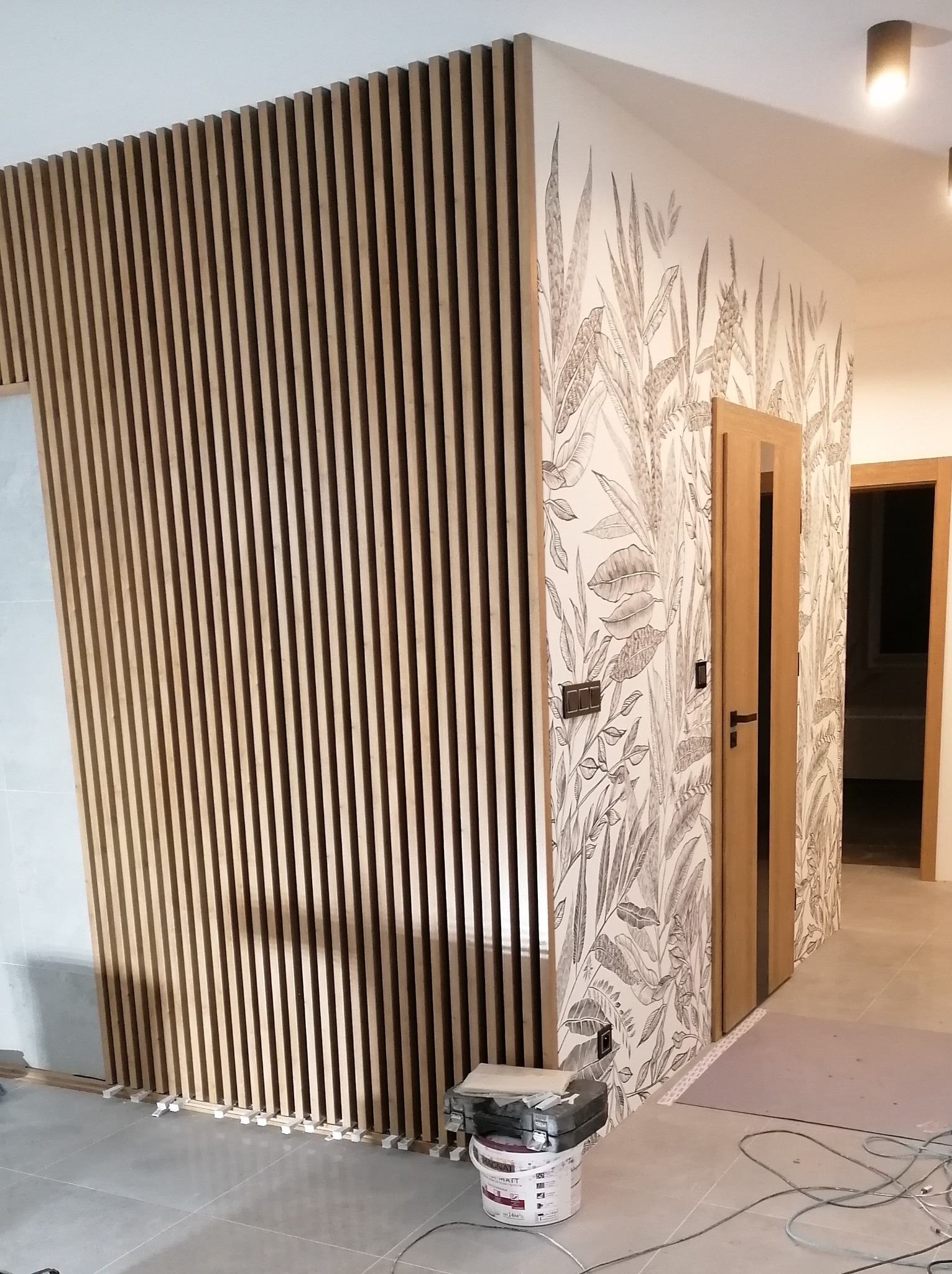 Montaż drewnianych lameli ściennych w przedpokoju z tapetą w liście, widoczny fragment podłogi z płytkami i akcesoria montażowe.