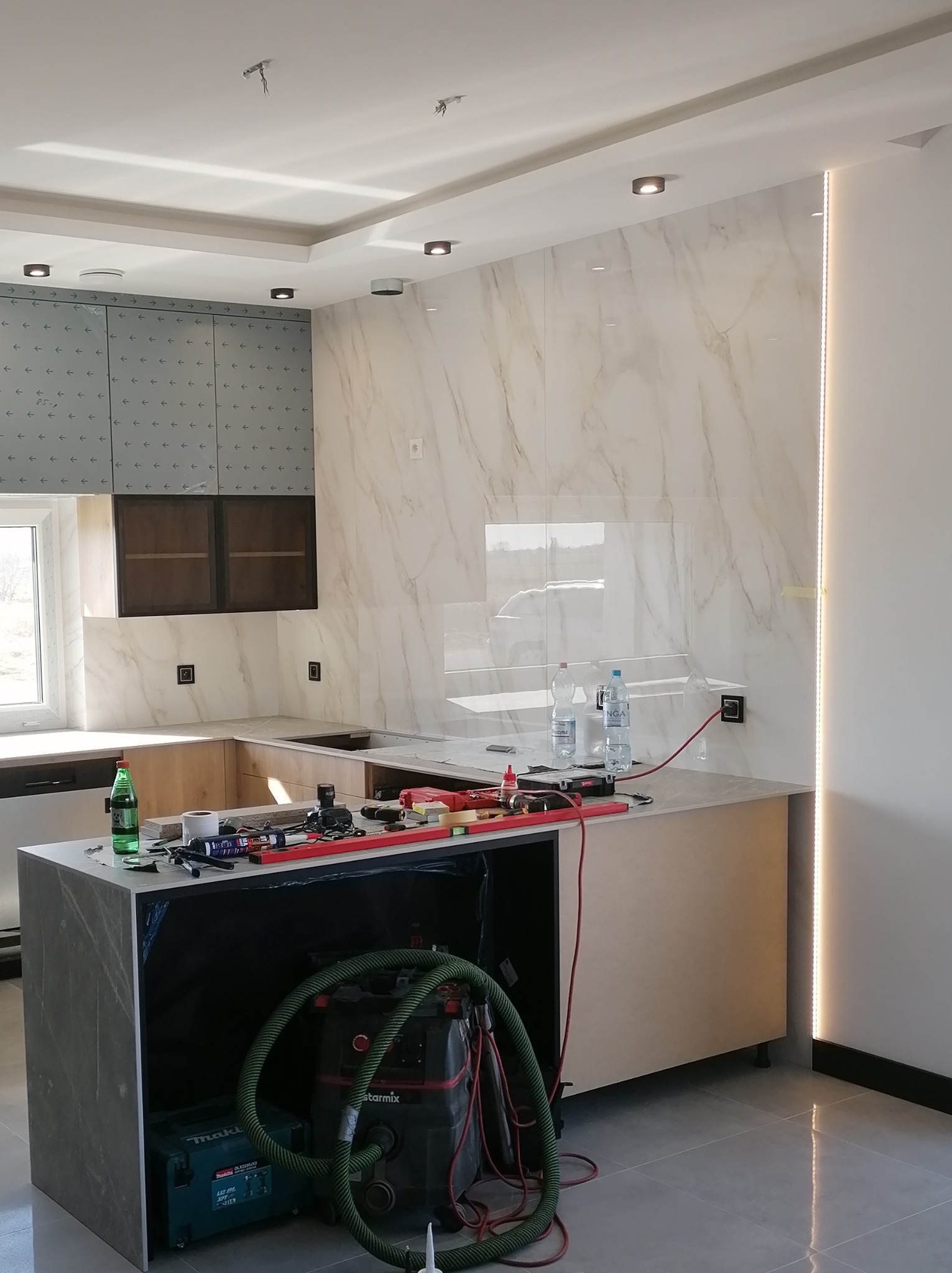 Wnętrze kuchni w trakcie prac wykończeniowych: marmurowe płytki na ścianie, częściowo zamontowane szafki z szarymi frontami, blat roboczy z narzędziami, odkurzacz przemysłowy i butelki z wodą...