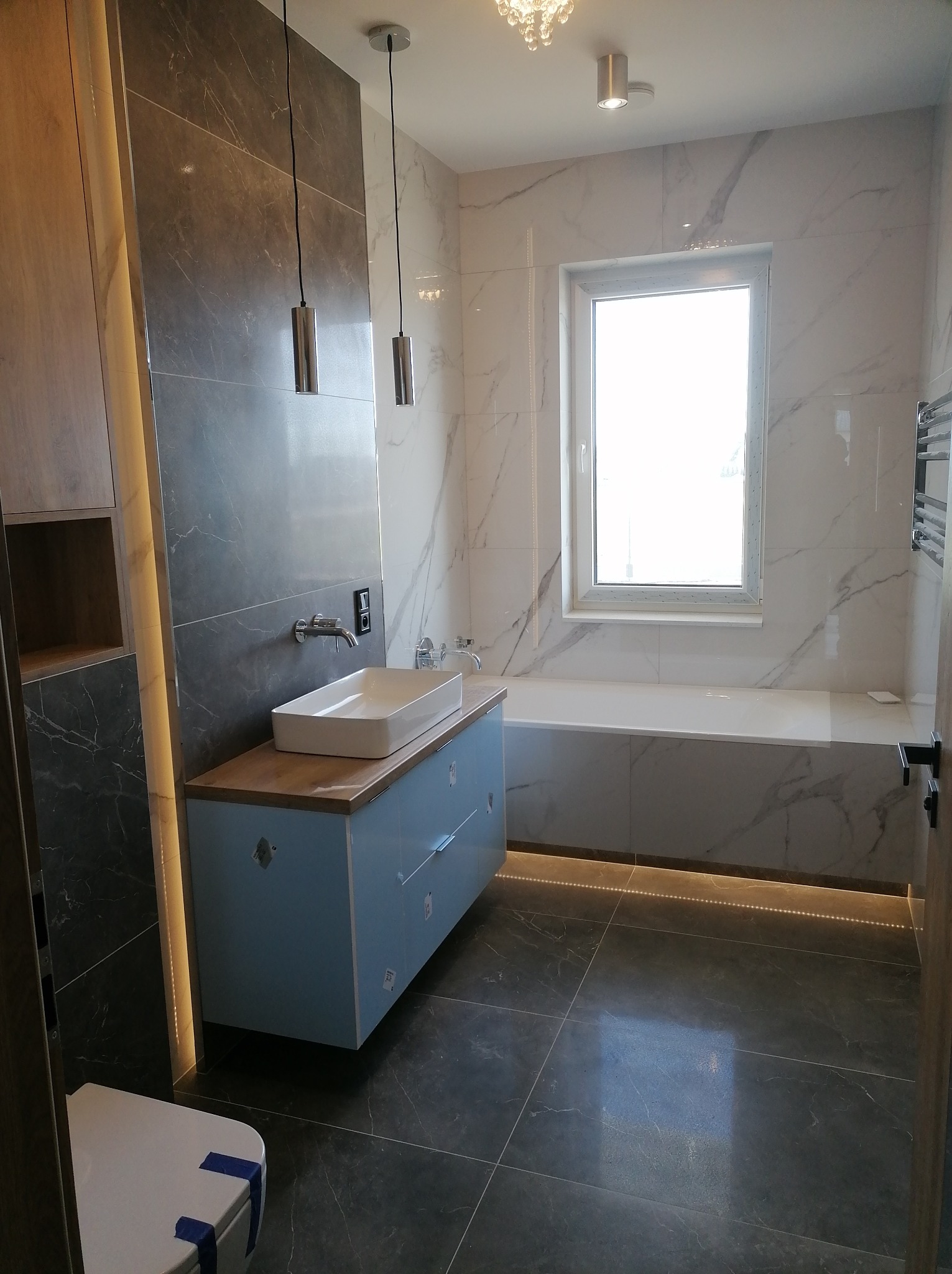 Nowoczesna łazienka z marmurowymi płytkami, podświetlaną wanną, błękitną szafką pod umywalką i wiszącymi lampami cylindrycznymi.